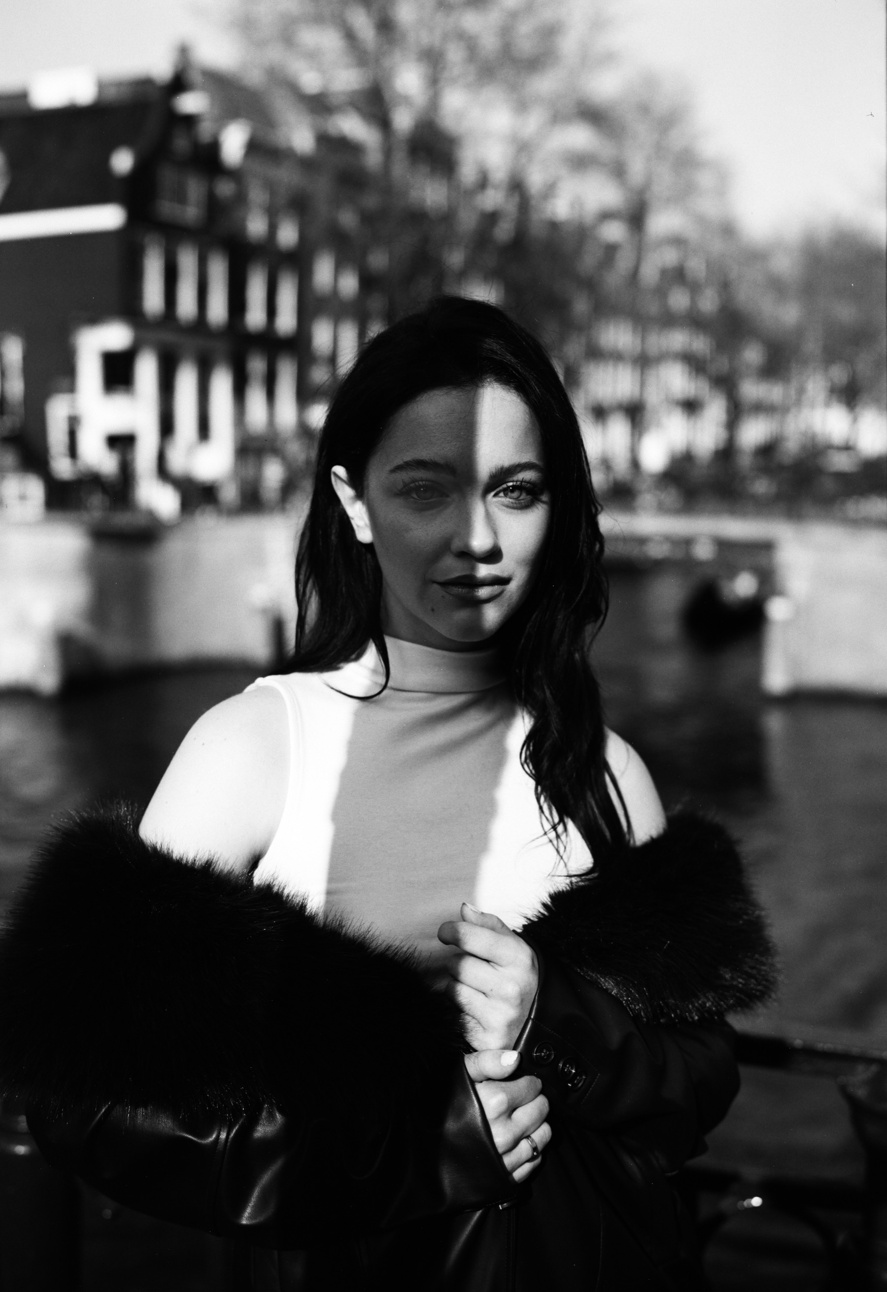 Zwart-witportret op Fomapan van een jonge vrouw aan de gracht, de zon over de helft van haar gezicht en een jas met bont om haar schouders