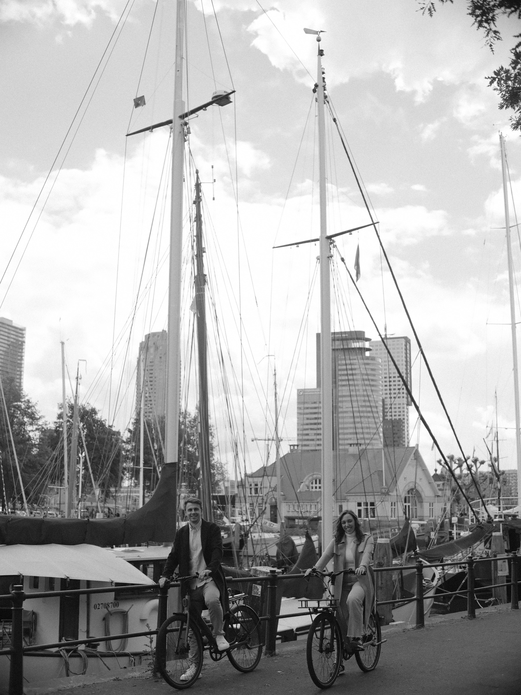 Black and white view of tall sailing ship masts in the Veerhaven, modern towers rising behind and a couple cycling past on the quay below