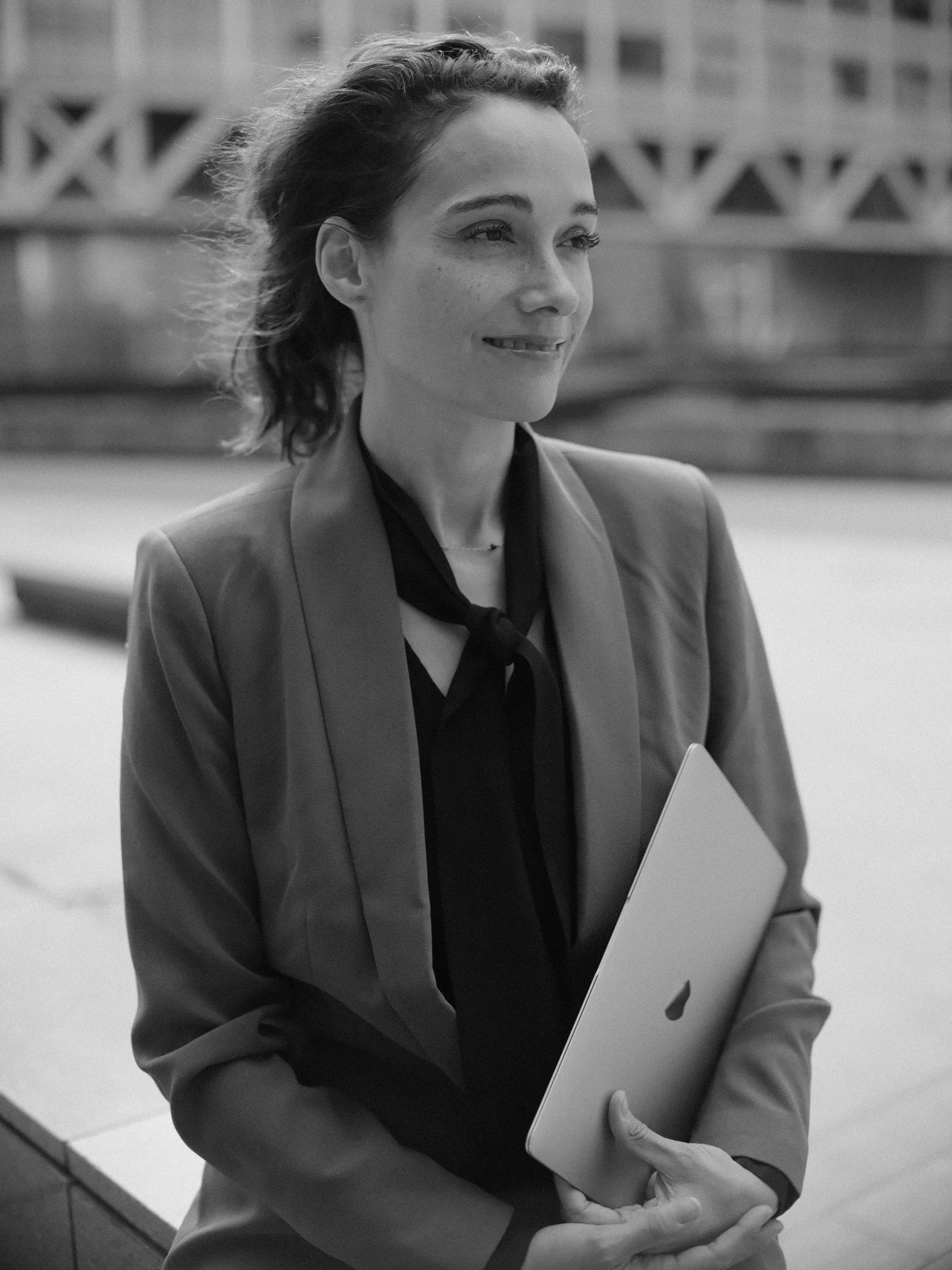 Vrouw in een colbert en een zwarte strikblouse kijkt glimlachend opzij met een laptop in haar handen, tijdens een solo fotoshoot in Den Haag