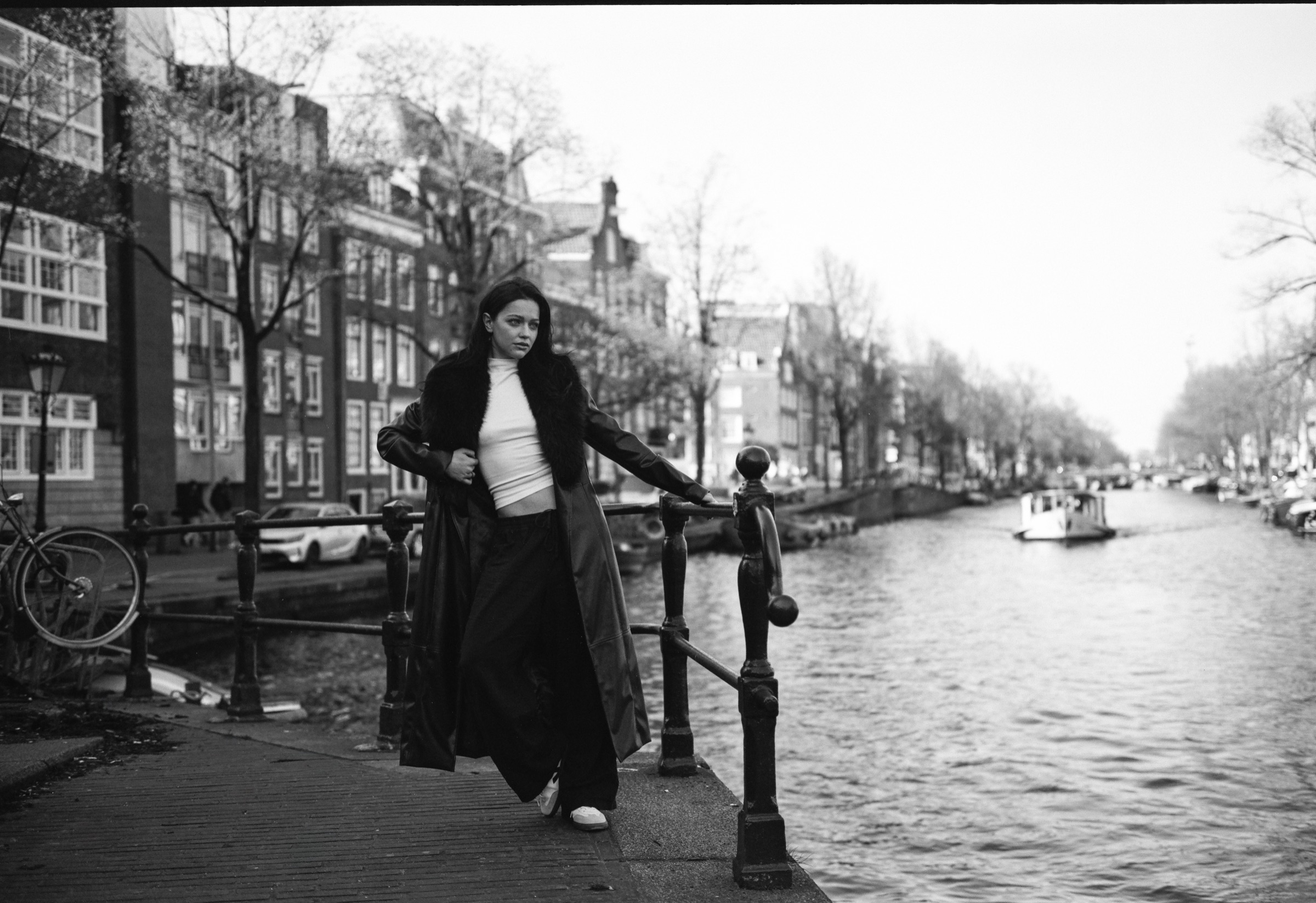 Vrouw in een lange donkere jas leunt tegen de ijzeren paaltjes langs een Amsterdamse gracht, terwijl er een bootje voorbijvaart