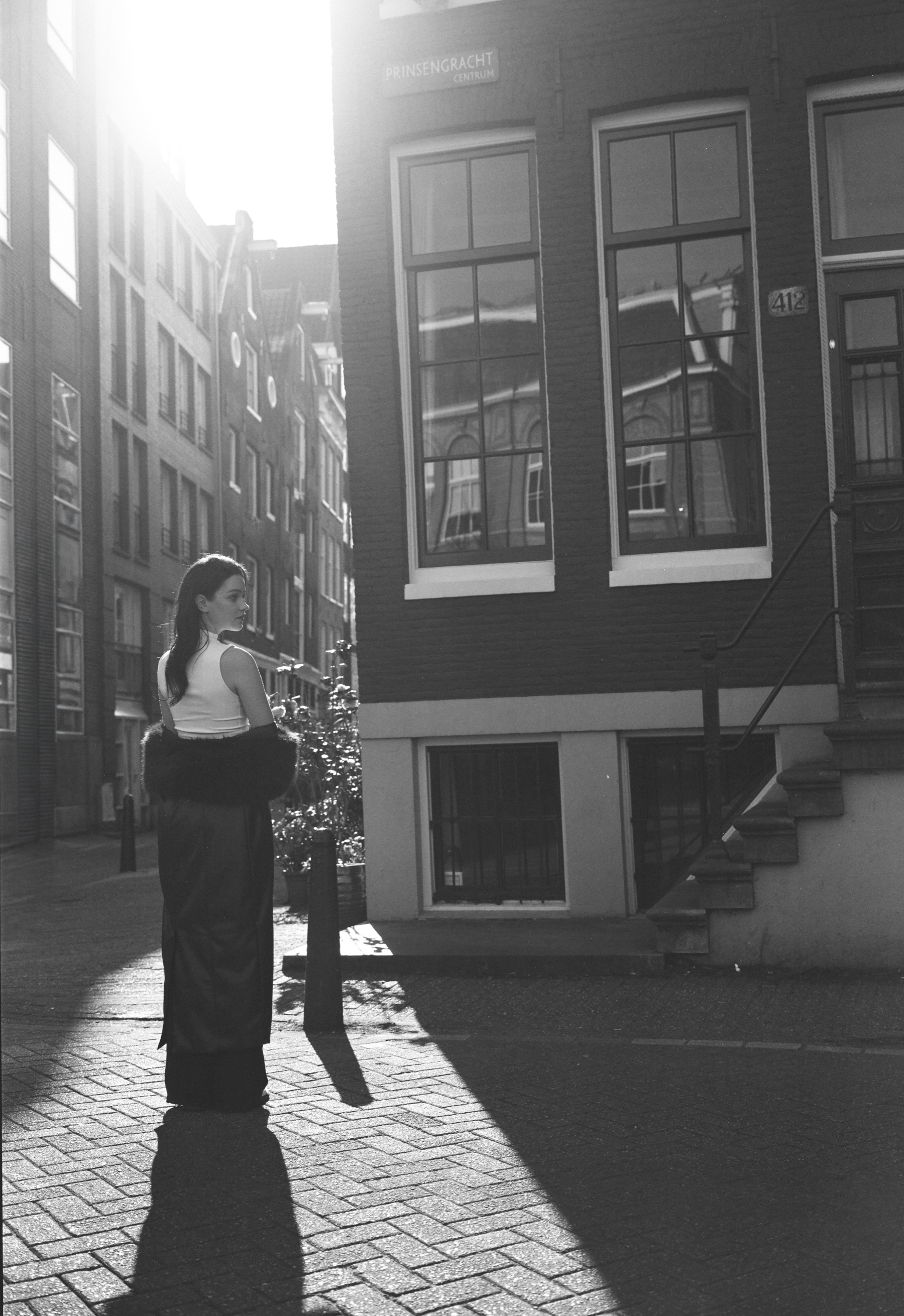 Vrouw in een wit topje en een wijde broek bij een hoek in de grachtengordel, ze kijkt over haar schouder in het felle zonlicht