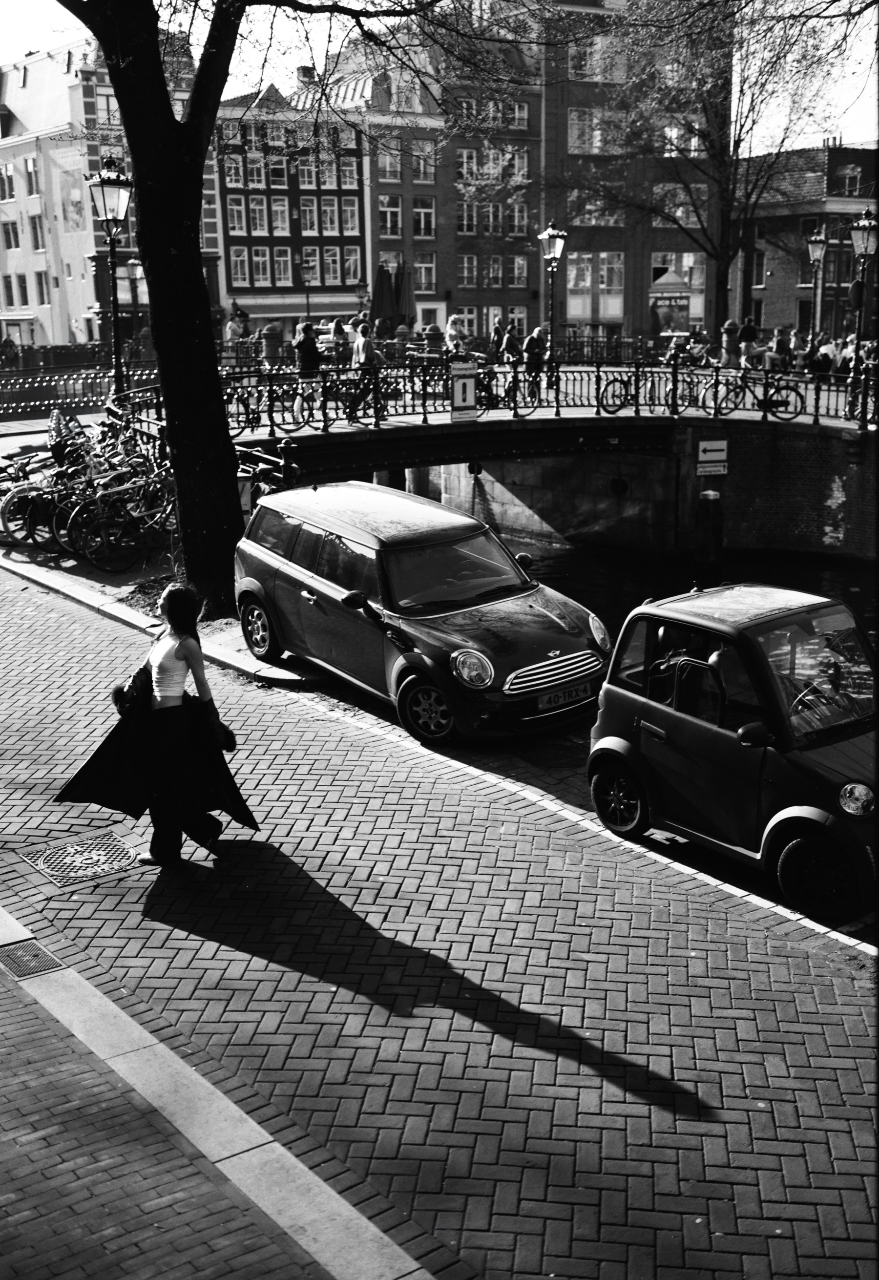 Vrouw loopt langs geparkeerde auto’s in een zonnige grachtenstraat, haar jas wappert en haar schaduw valt lang over de klinkers