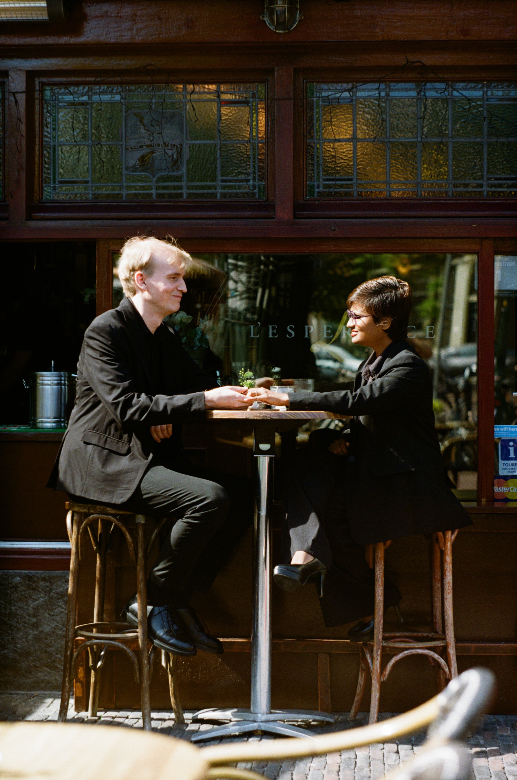 Couple holding hands across a small table on tall wooden stools outside a café with stained glass, sun on their black clothes