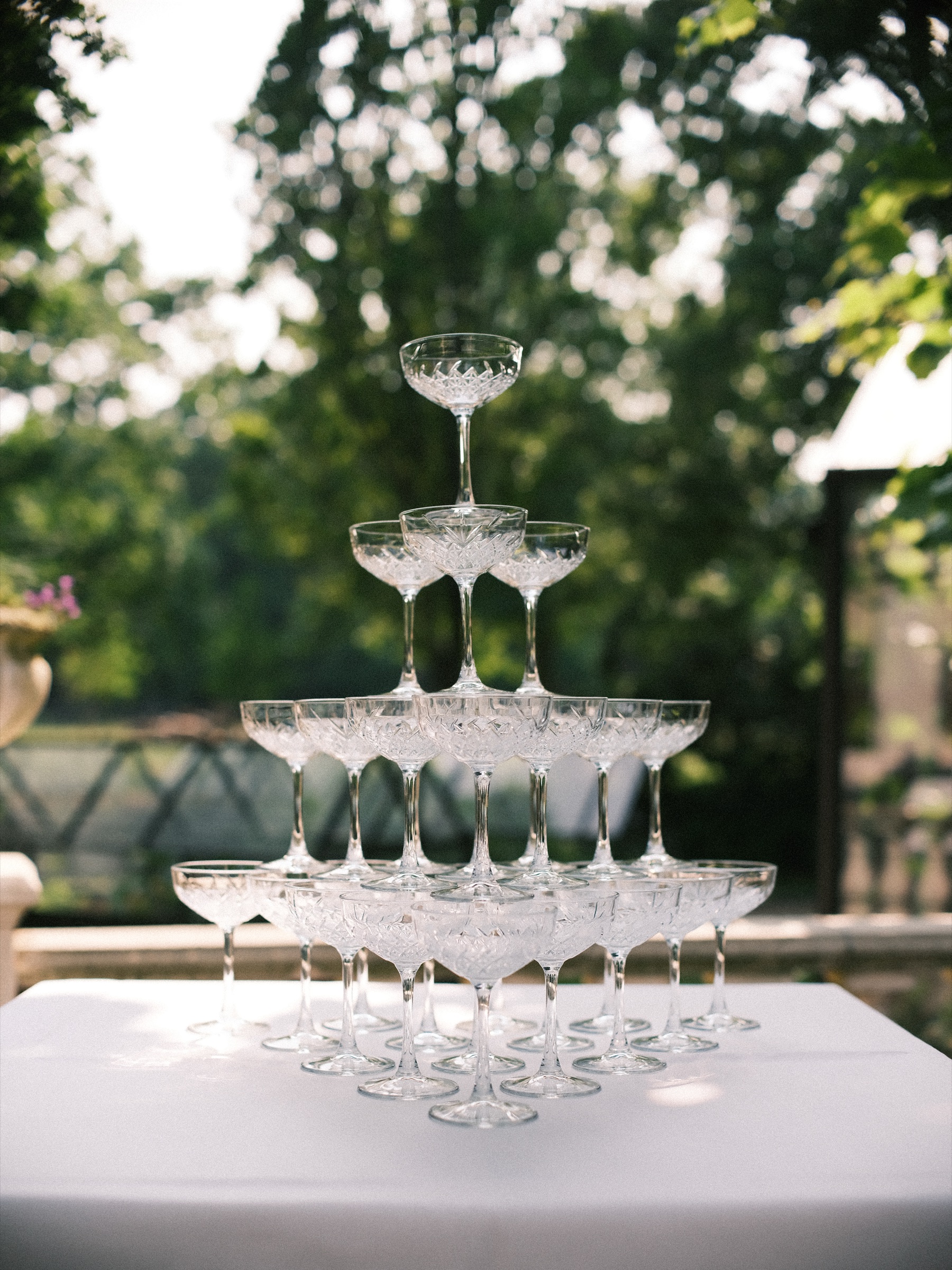 Toren van lege champagneglazen op een witte tafel in de tuin, met groene bomen en glinsterend water wazig op de achtergrond