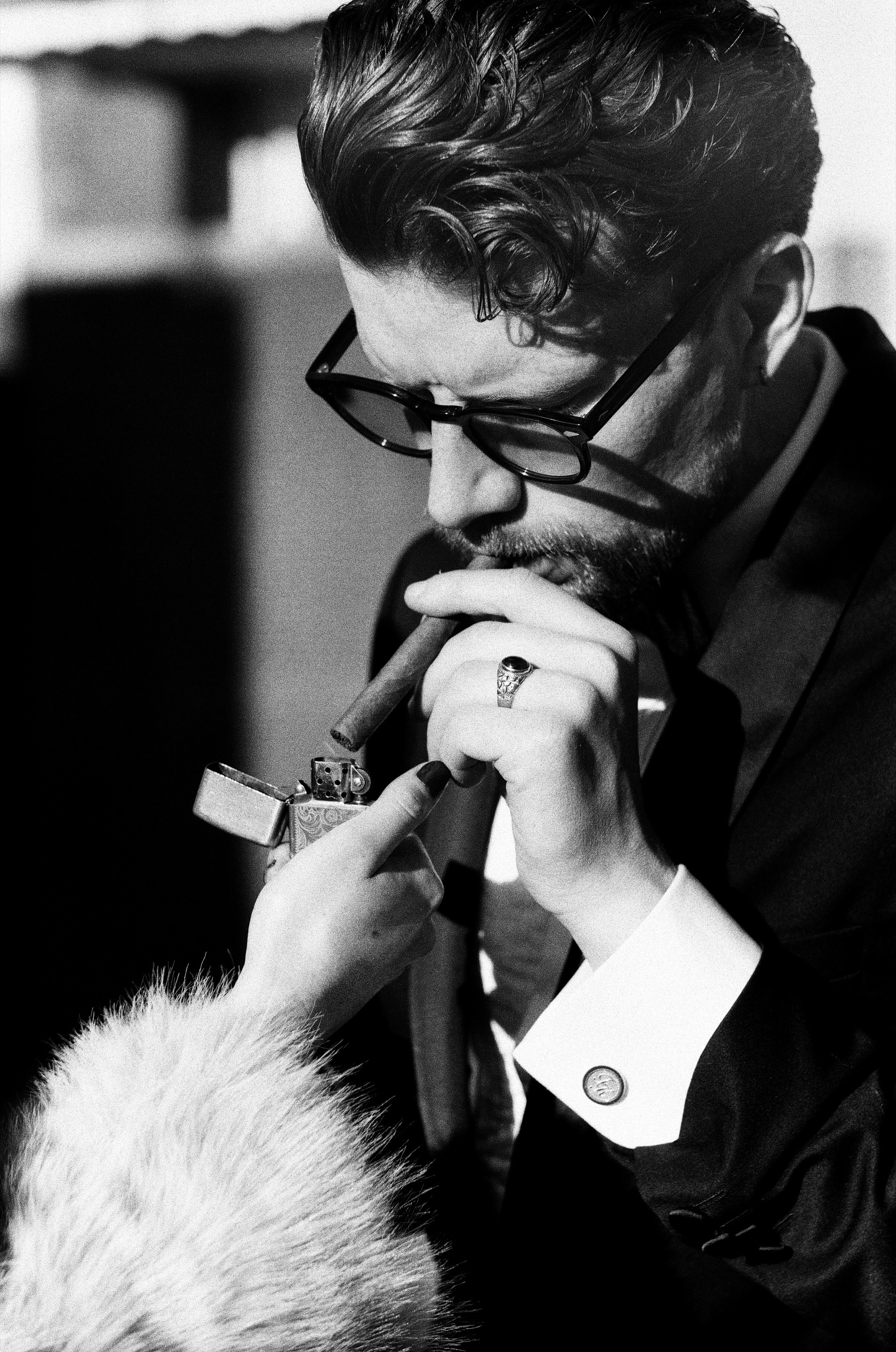 Close view of him lighting a cigar from a metal lighter she holds, her fur sleeve in the frame and his cufflink catching the sun
