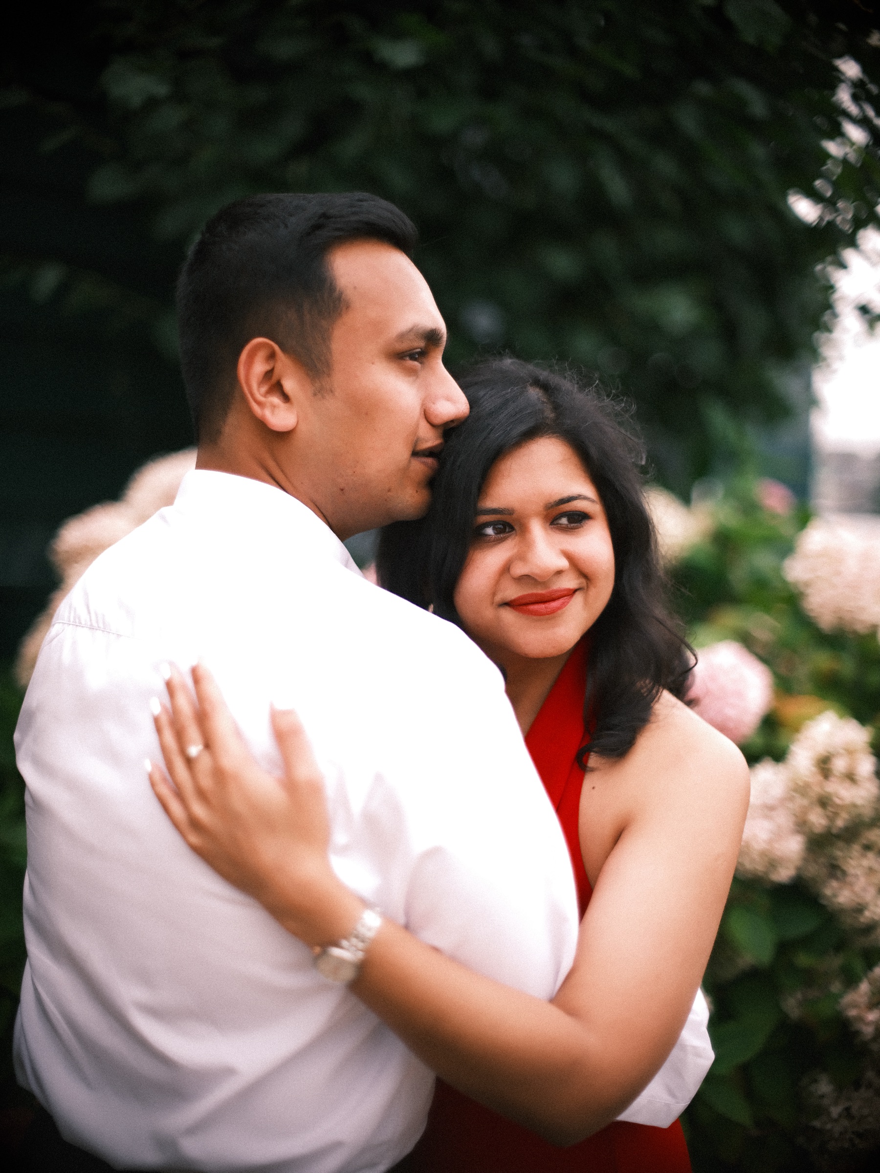 Close embrace by cream and pink hydrangeas, her eyes on the lens over his shoulder and her hand resting on his white shirt