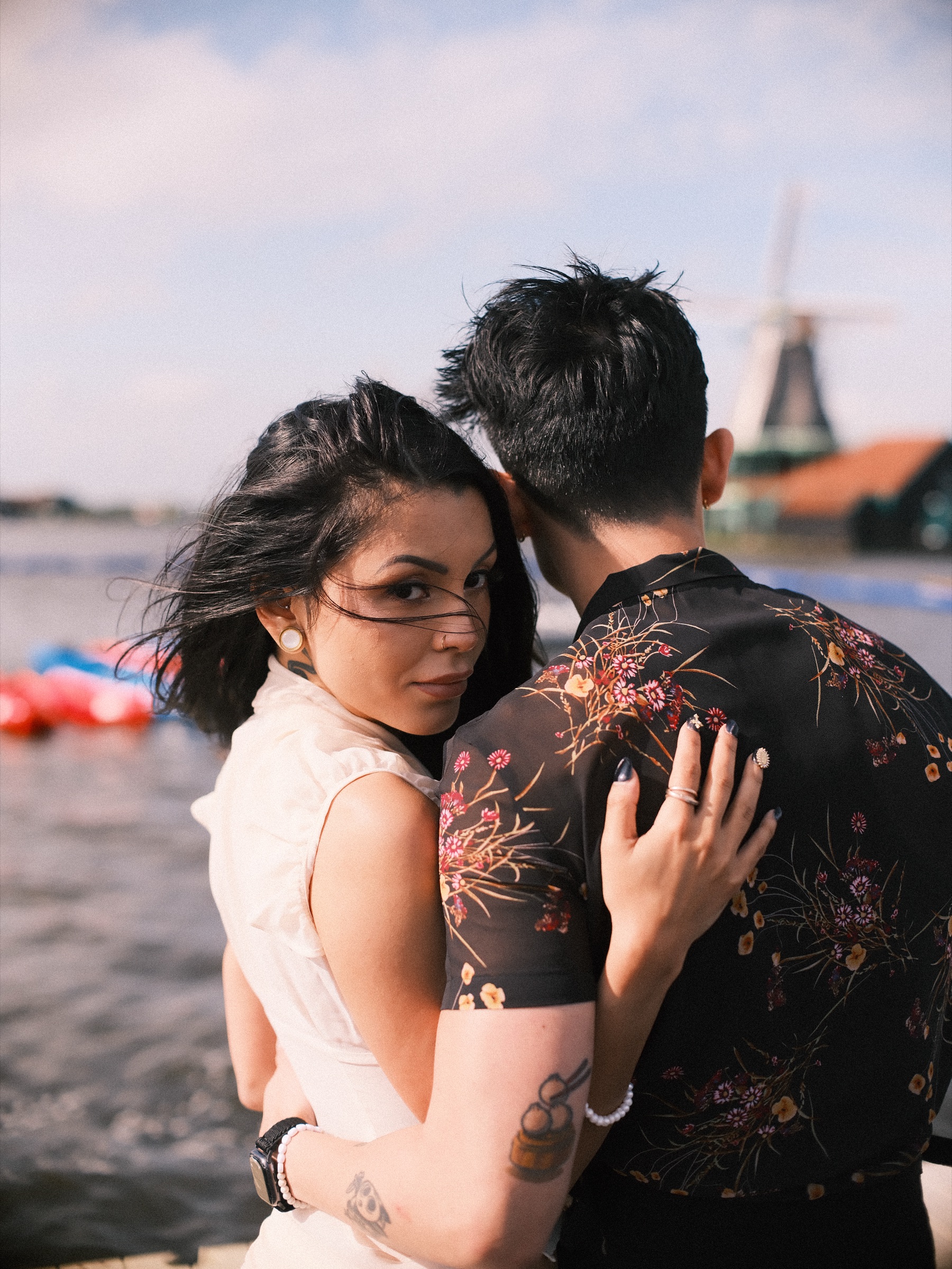 Close embrace by the water, her windblown hair across her face as she looks over his shoulder, his floral shirt in sharp focus