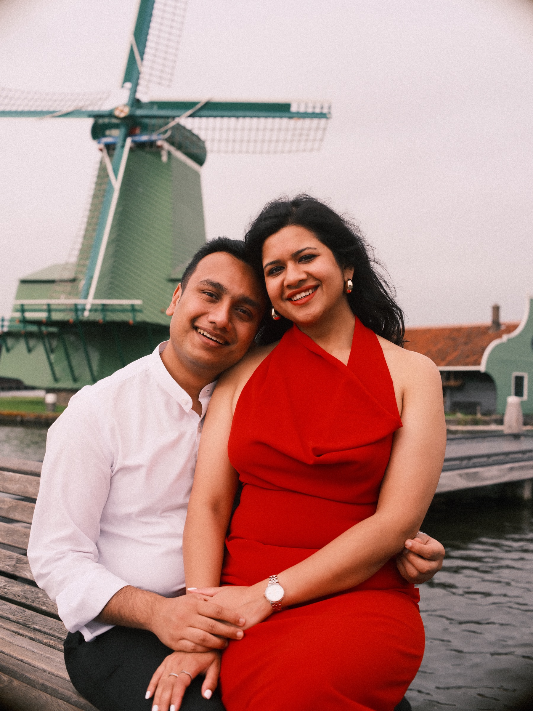 Close portrait on the bench, her head resting against his as both smile, the green windmill and orange roofs behind them