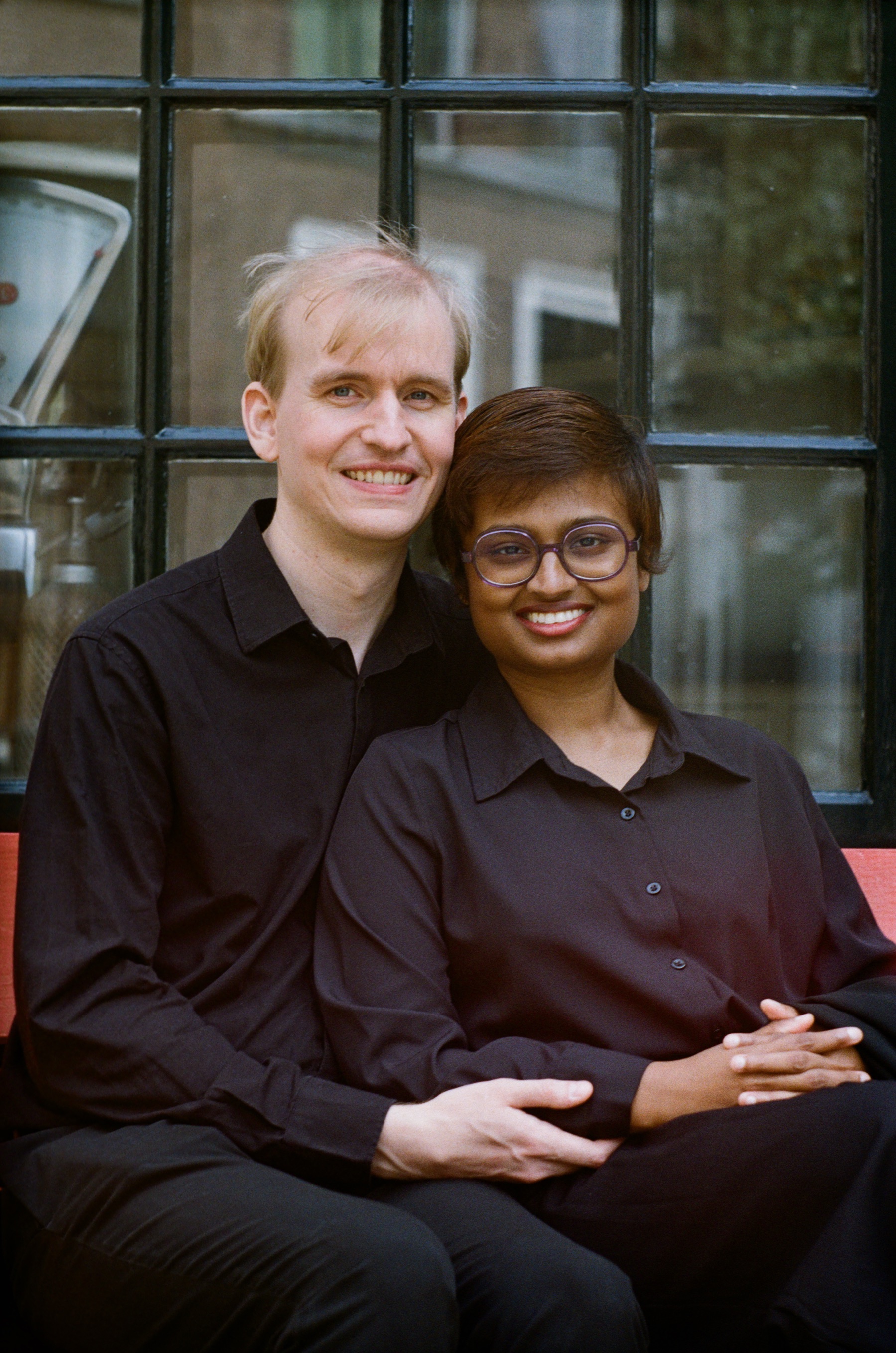 Close portrait of a smiling couple sitting in front of a dark window, his arm around her and her hands folded on her knee