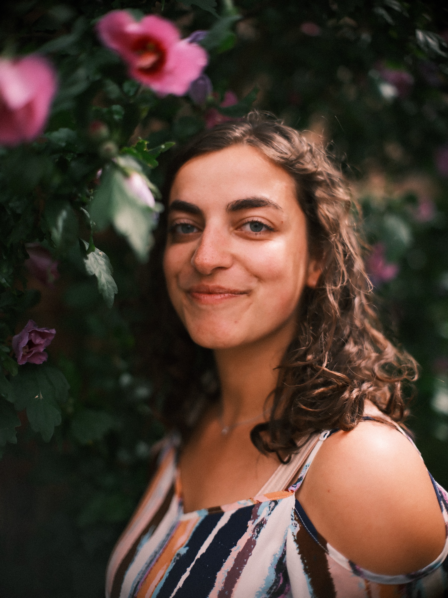 Portret van dichtbij van een vrouw met krullen, een roze bloem boven haar en donker blad erachter, zomer in Haarlem