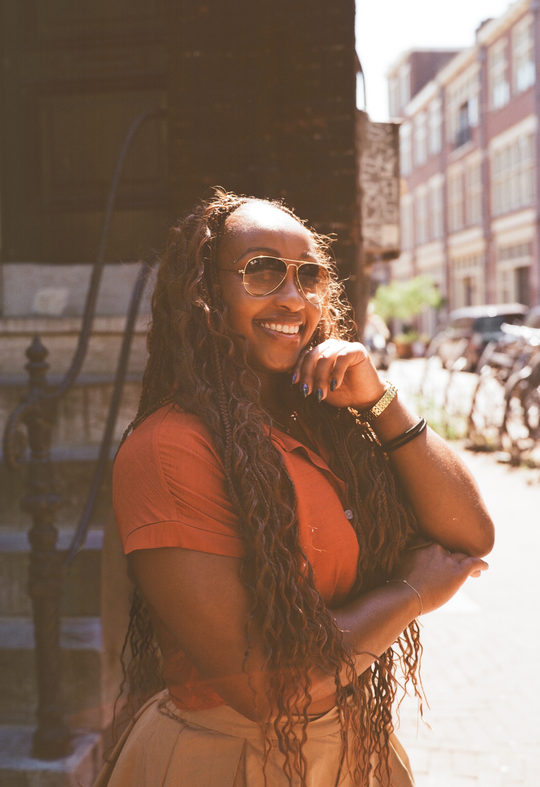 Close portrait in warm sun of a smiling woman in sunglasses resting her chin on her hand, long braids over an orange shirt