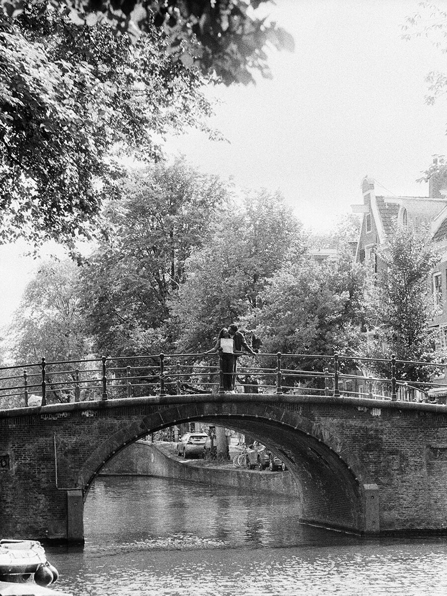 Wide analog portrait of a couple standing on an arched canal bridge beneath leafy trees in Amsterdam