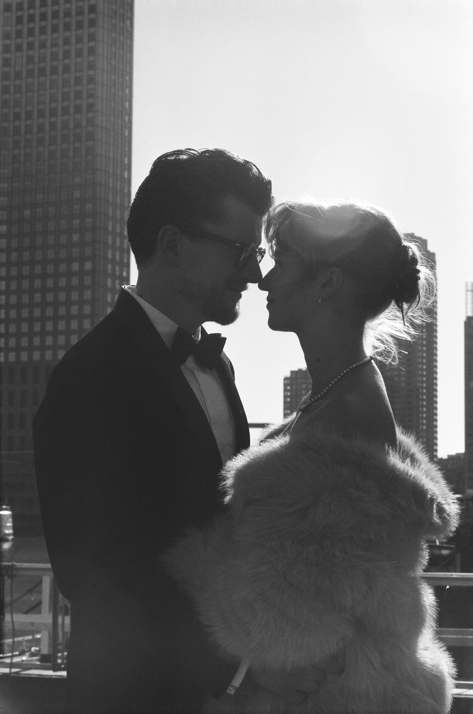 Backlit and almost nose to nose, the couple stands out against tall towers, a halo of sun around her pinned-up hair
