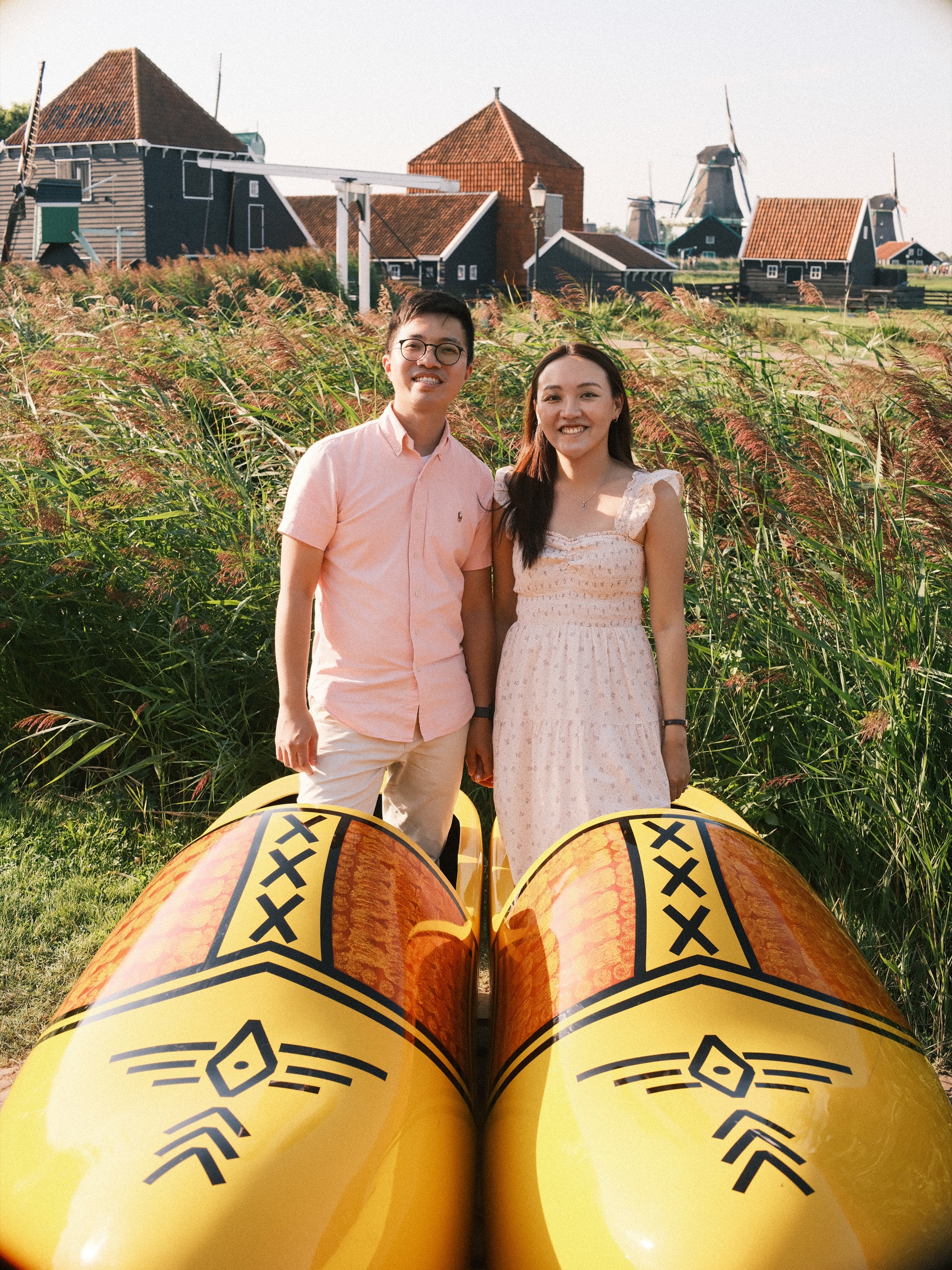 Couple standing behind two giant yellow clogs in tall reeds, red-roofed houses and windmills of the Zaanse Schans behind them