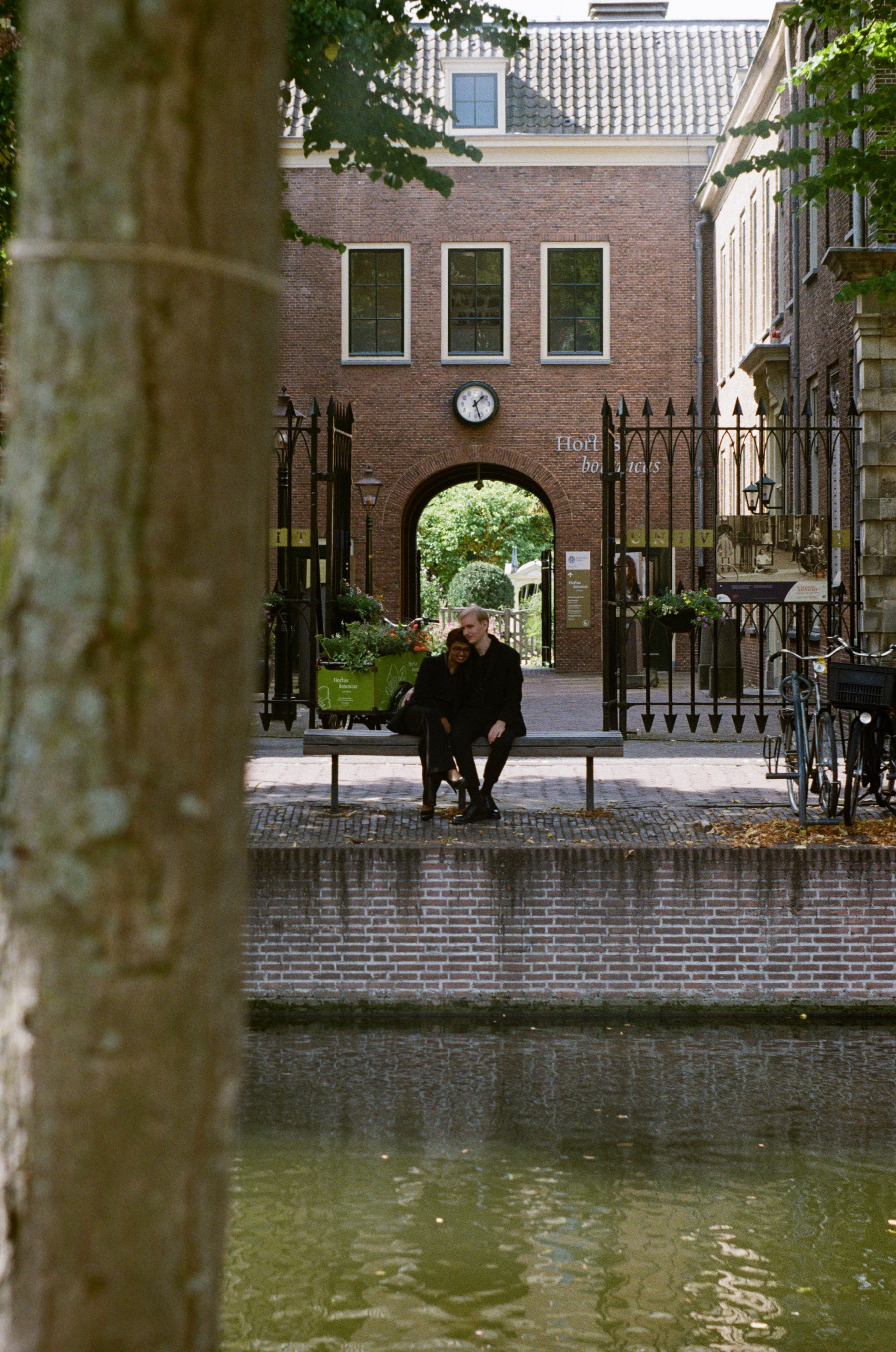 Couple on a bench before the Hortus botanicus gate, seen across the Rapenburg past a tree trunk, photos in Leiden on a roll of Kodak Gold