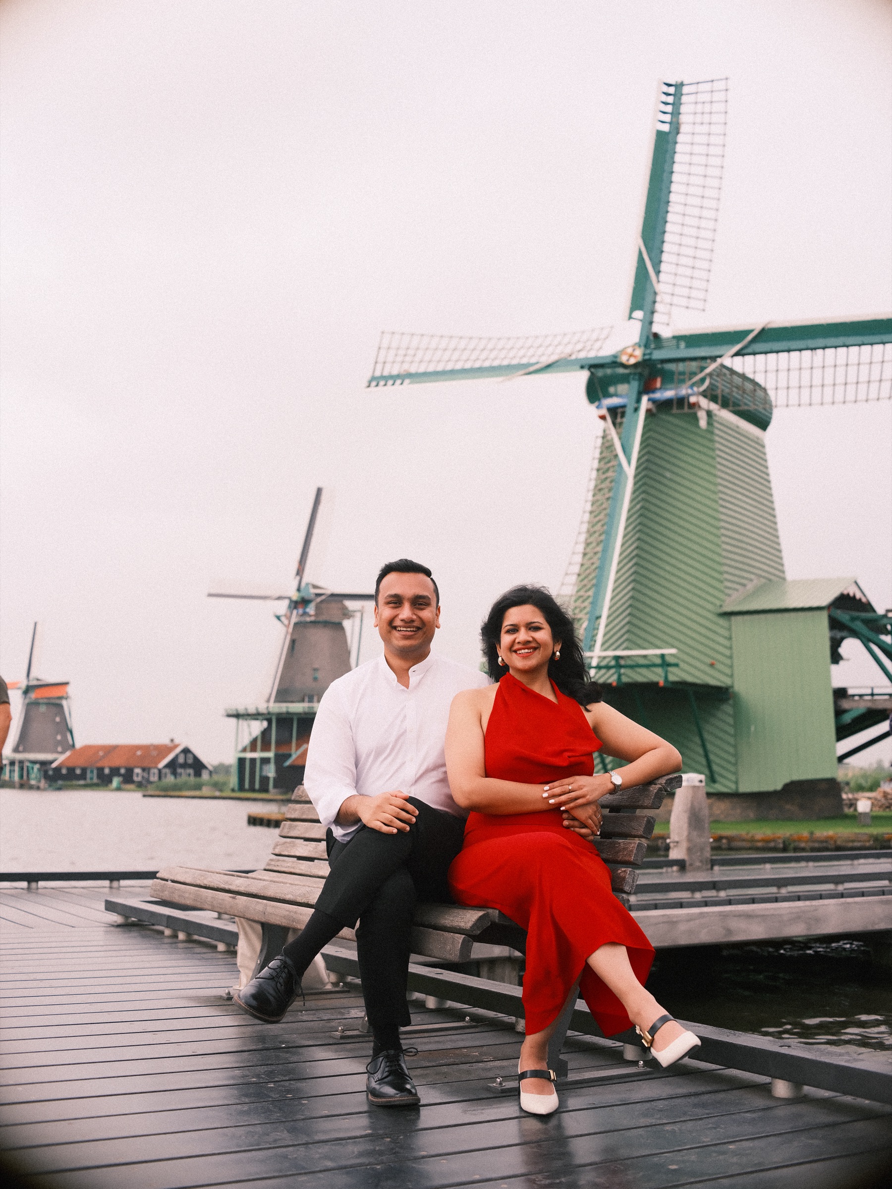 Smiling couple on a wooden bench on a jetty at the Zaanse Schans, she in a red dress, the big green mill rising behind them