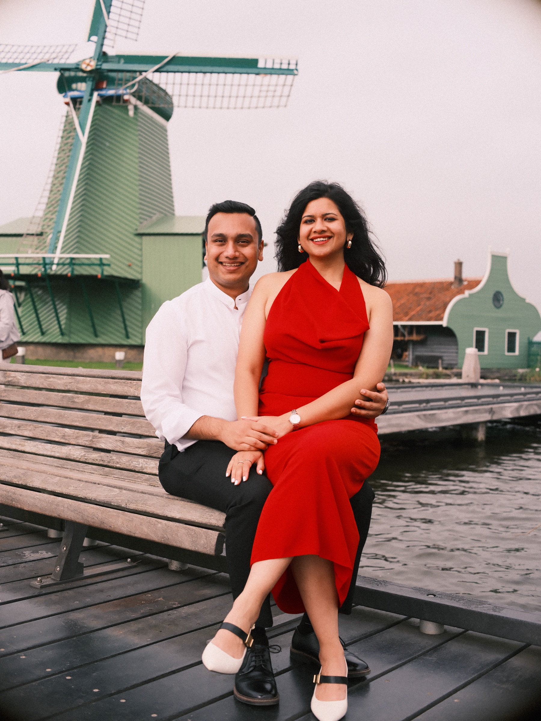 Couple sitting on a wooden bench on the jetty, she in a red dress with white shoes, the big green windmill rising behind them