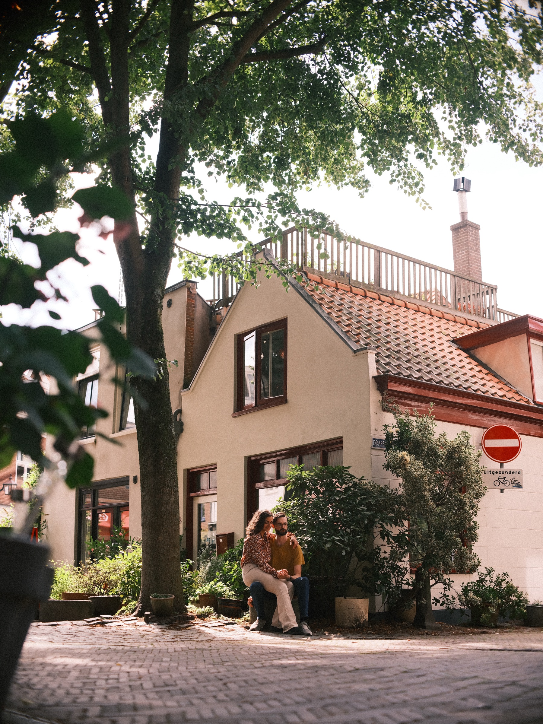 Stel zit dicht tegen elkaar onder een hoge boom op de Vijfhoek in Haarlem, met een crèmekleurig huis, een pannendak en een verbodsbord