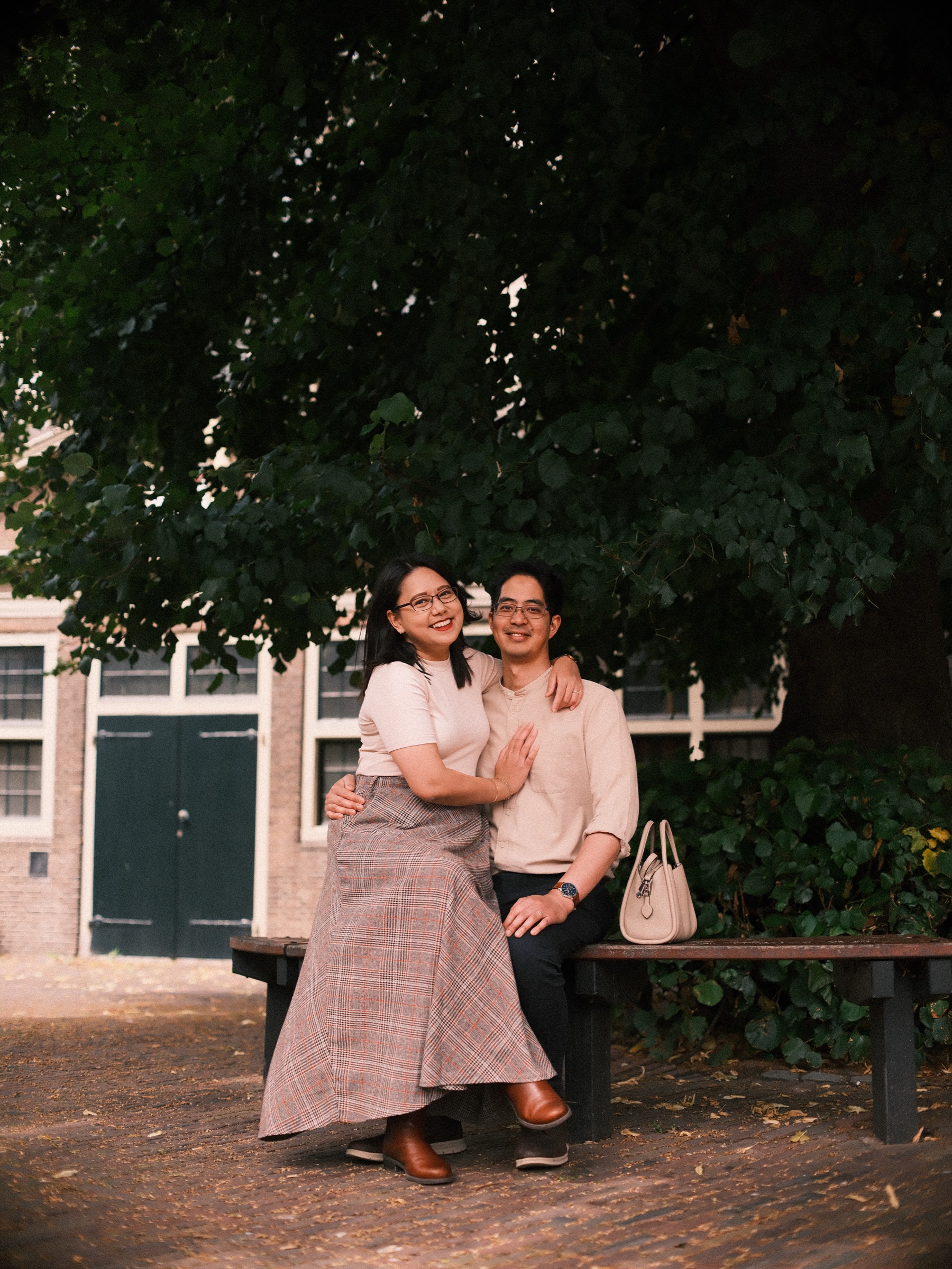 Couple laughing on a wooden bench in the deep shade of the broad tree on the Pieterskerkhof, small brick houses with green doors behind