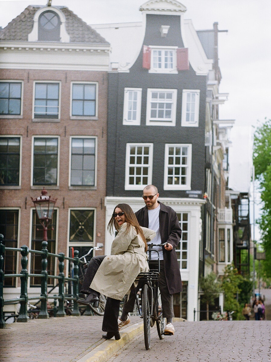 Laughing on a bicycle’s front rack, a woman poses with her partner during an analog couple photoshoot on an Amsterdam bridge