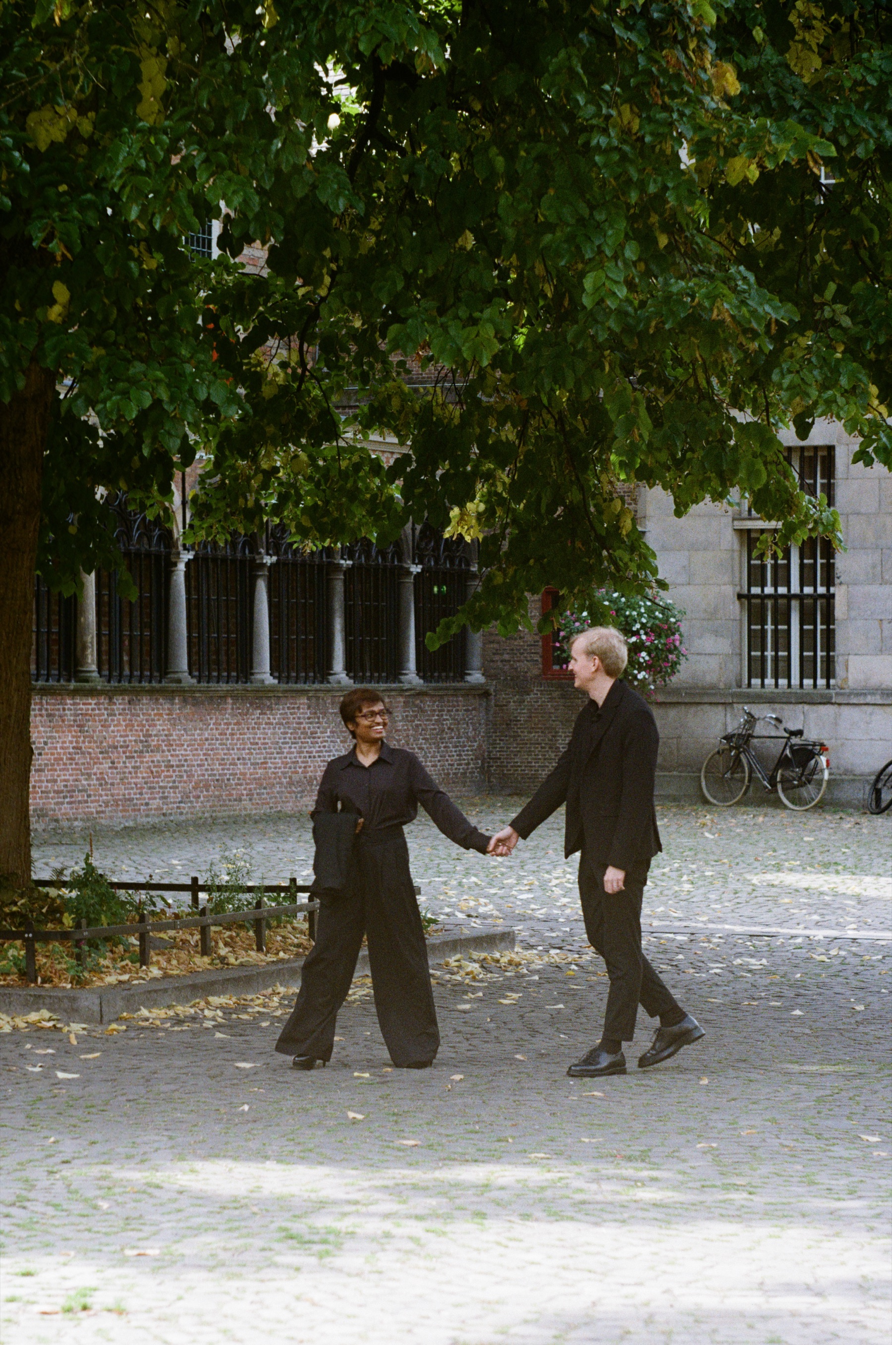 Couple in black holding hands on a sunlit cobbled square, stepping toward each other under a tree, fallen leaves on the stones