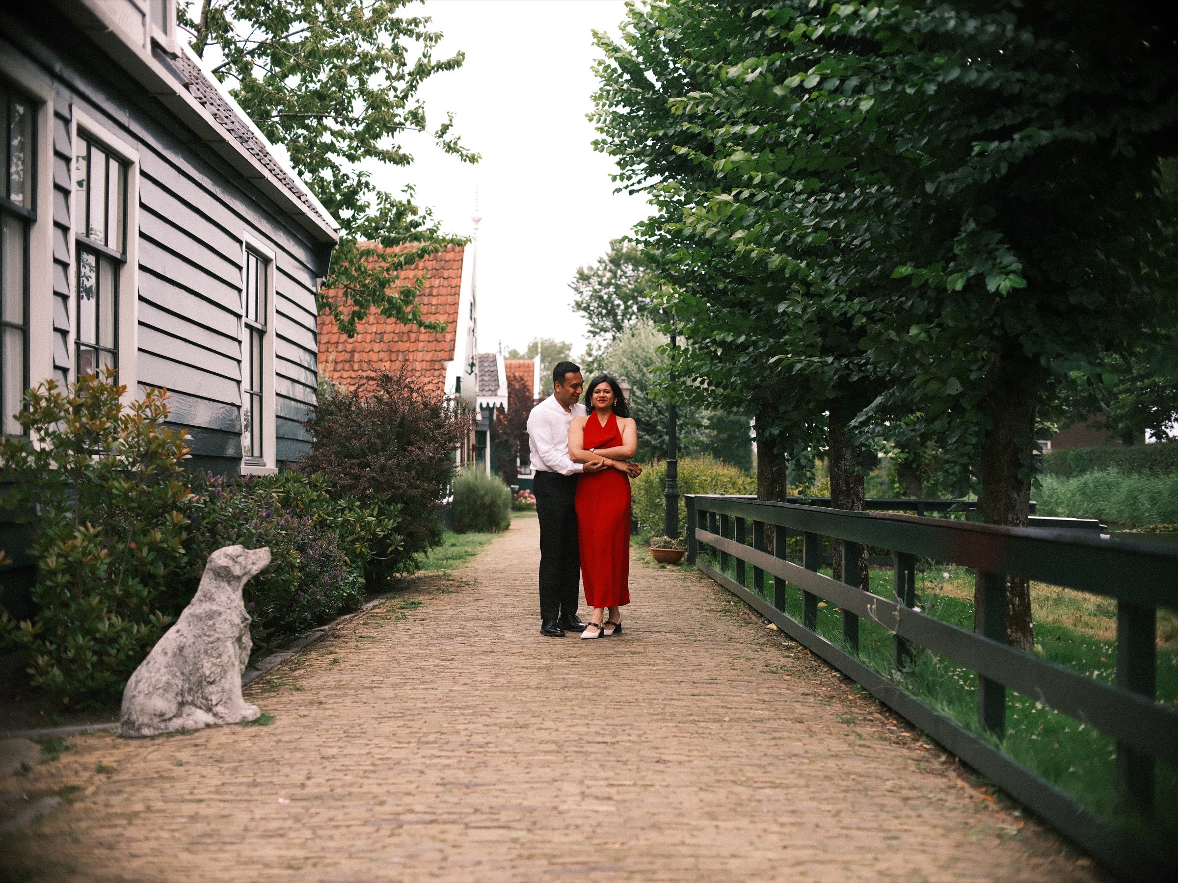 Man in a white shirt and woman in a red halter dress standing in a quiet brick lane at the Zaanse Schans, a stone dog statue in front