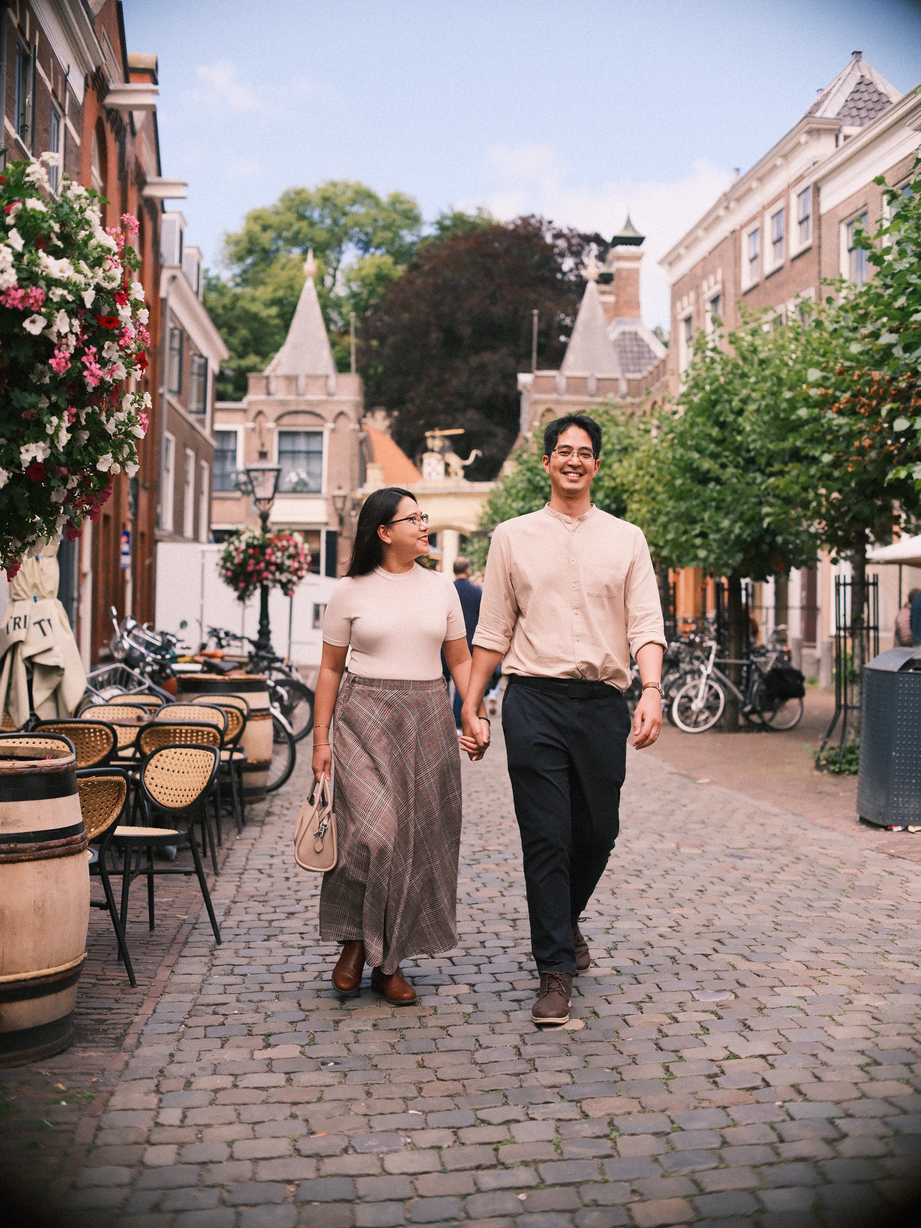 Couple walking hand in hand down a cobbled café lane toward the Burcht gate, a photo spot in Leiden with wine barrels and flower baskets