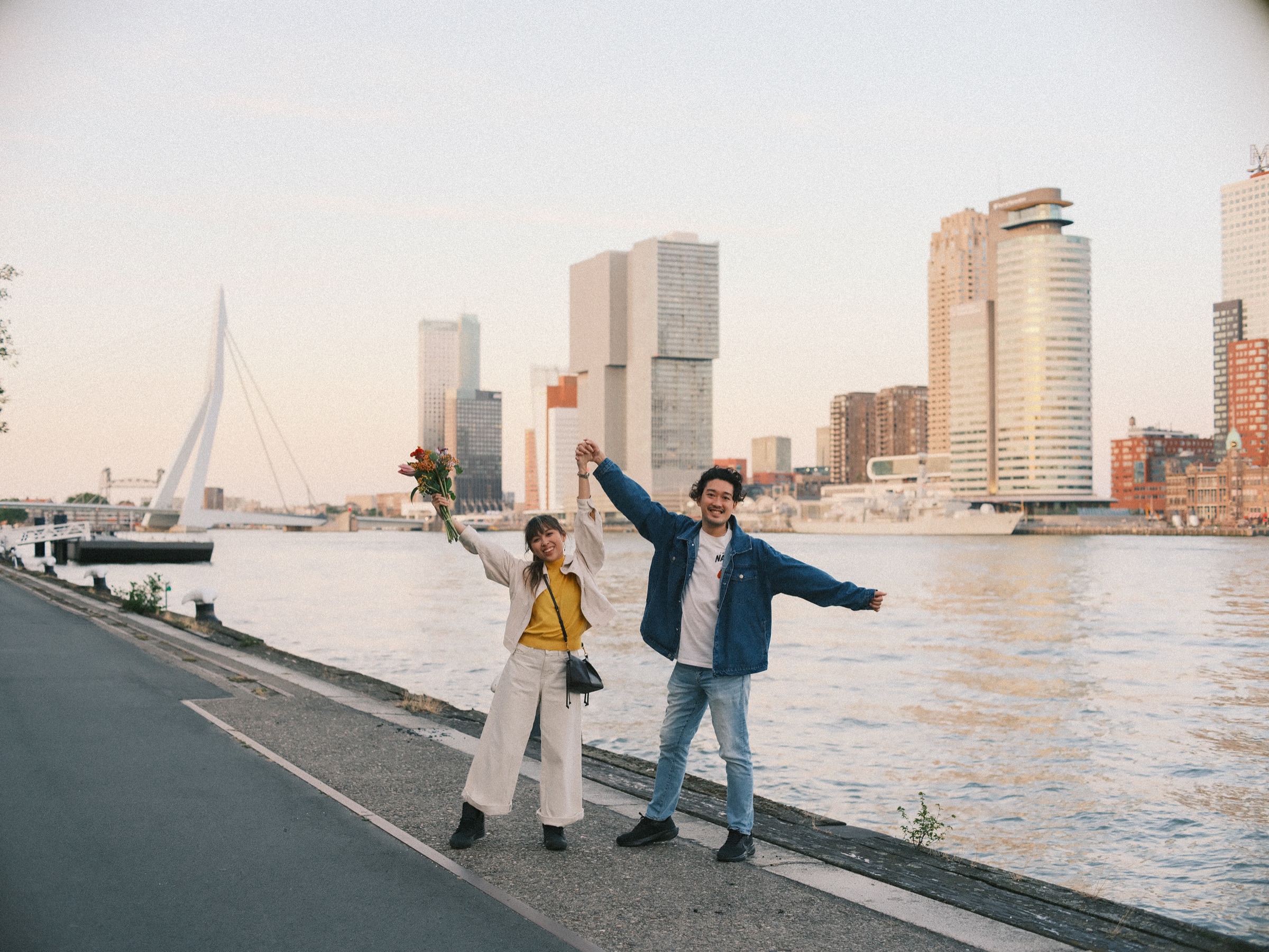 Stel juicht met de armen omhoog op de Maaskade, met de Erasmusbrug en de torens van de Kop van Zuid aan de overkant