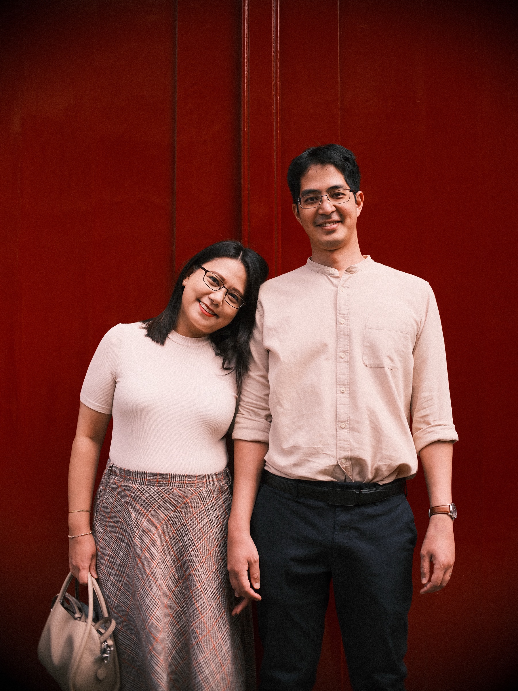 Couple leaning close against a deep red door, her head tilted toward his shoulder and both smiling while the red fills the whole frame