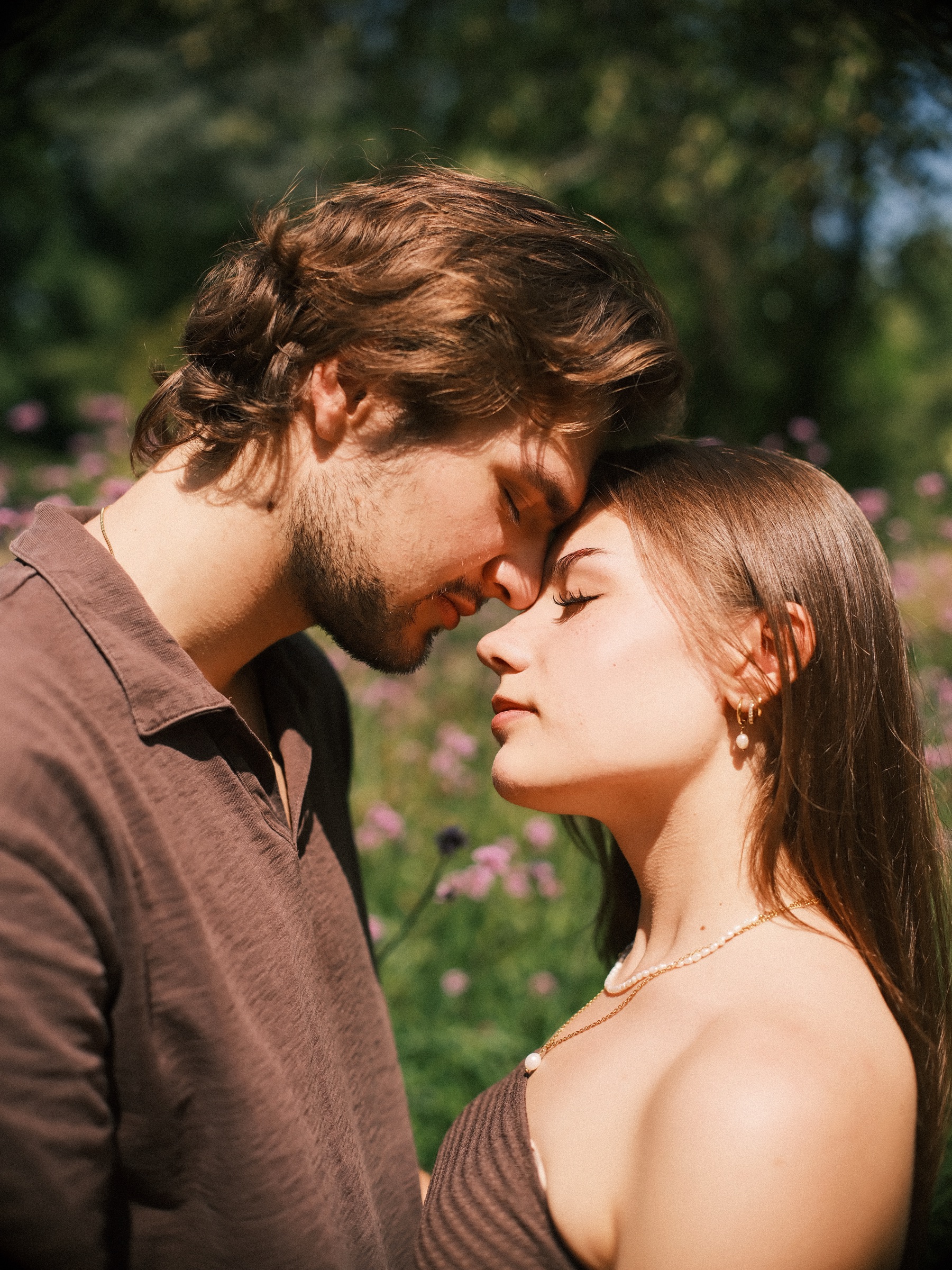 Close portrait of him resting his nose against her forehead as she closes her eyes, purple flowers and tall grass behind them