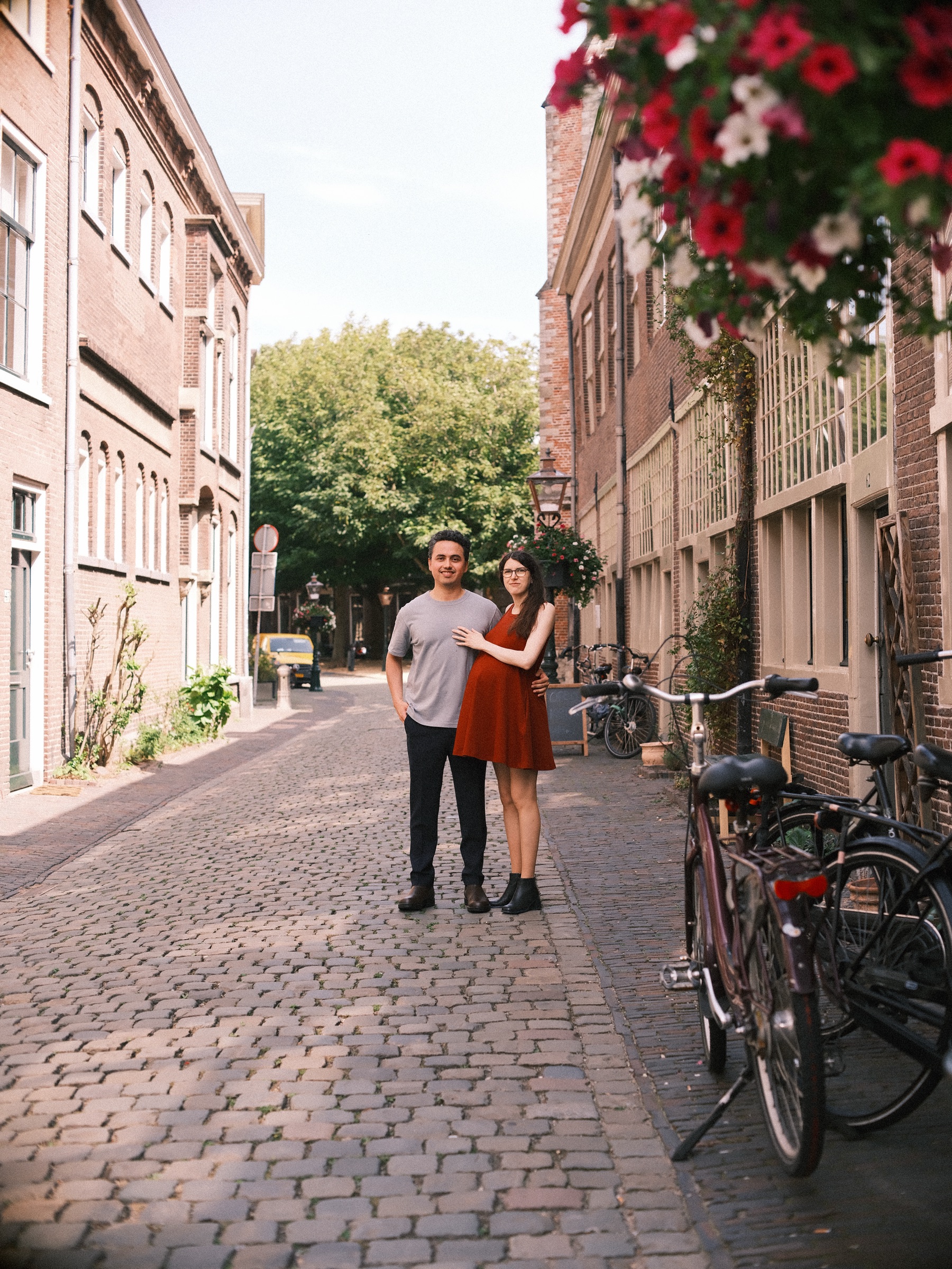 Couple standing far down a cobbled lane under a hanging basket of red flowers, bicycles parked along the brick houses
