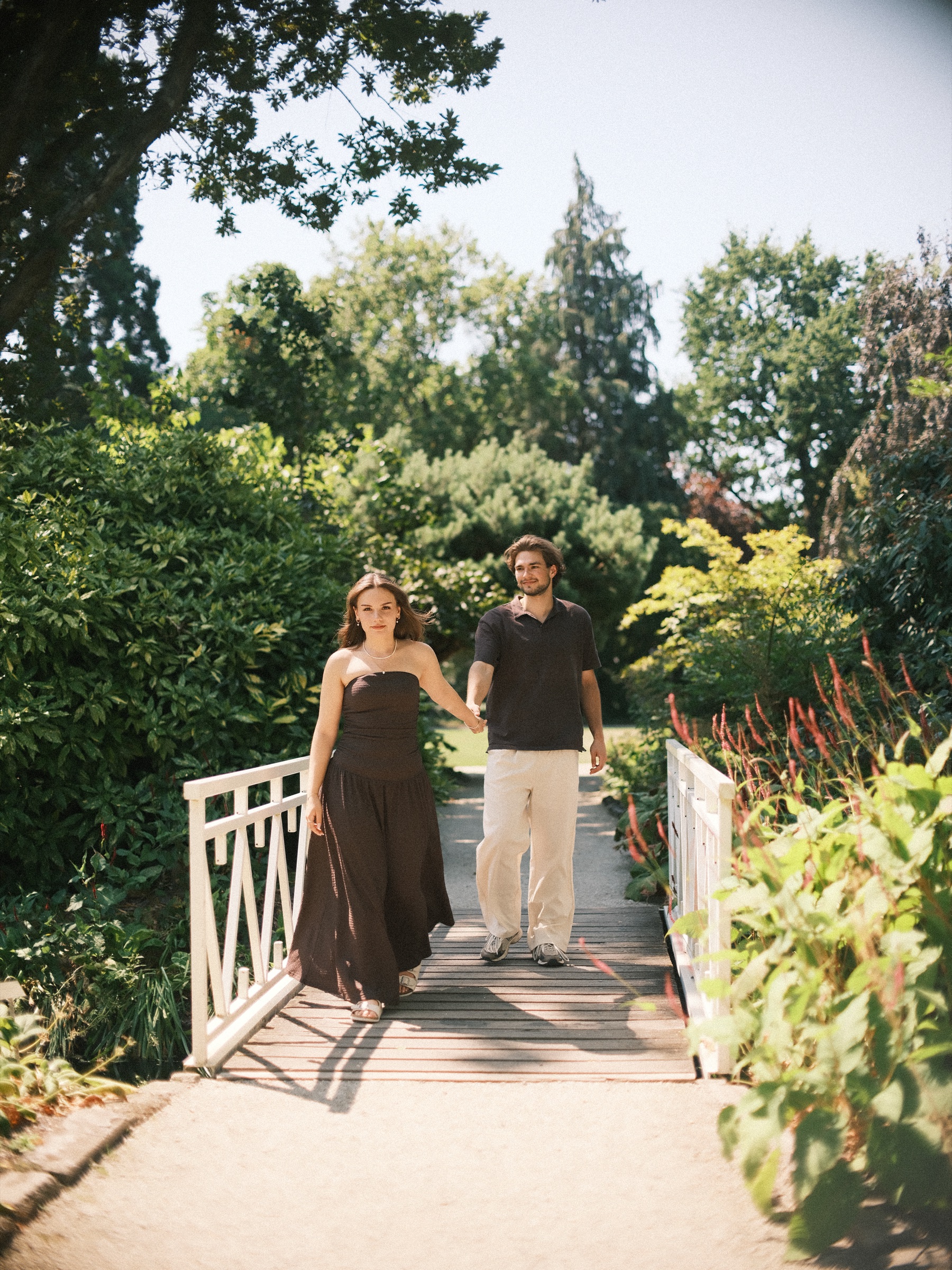 Couple crossing a small white wooden bridge lined with red flower spikes, tall trees behind, during a July photo session in Rotterdam