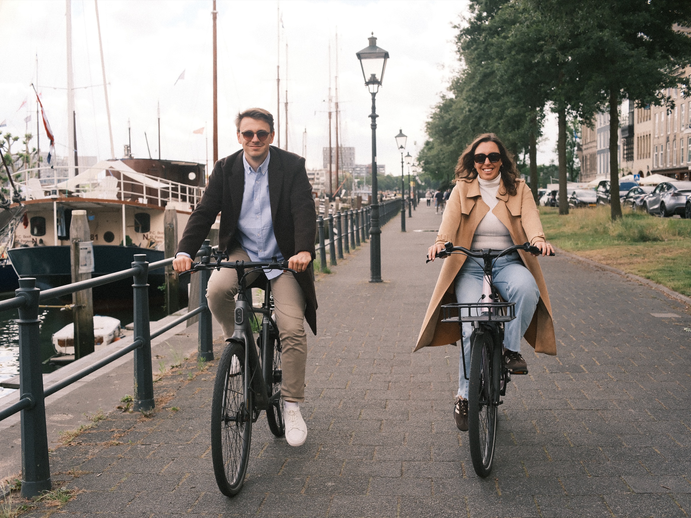 Cycling side by side toward the lens on a tree-lined quay with old lanterns, she in a camel trench coat and he in a dark coat