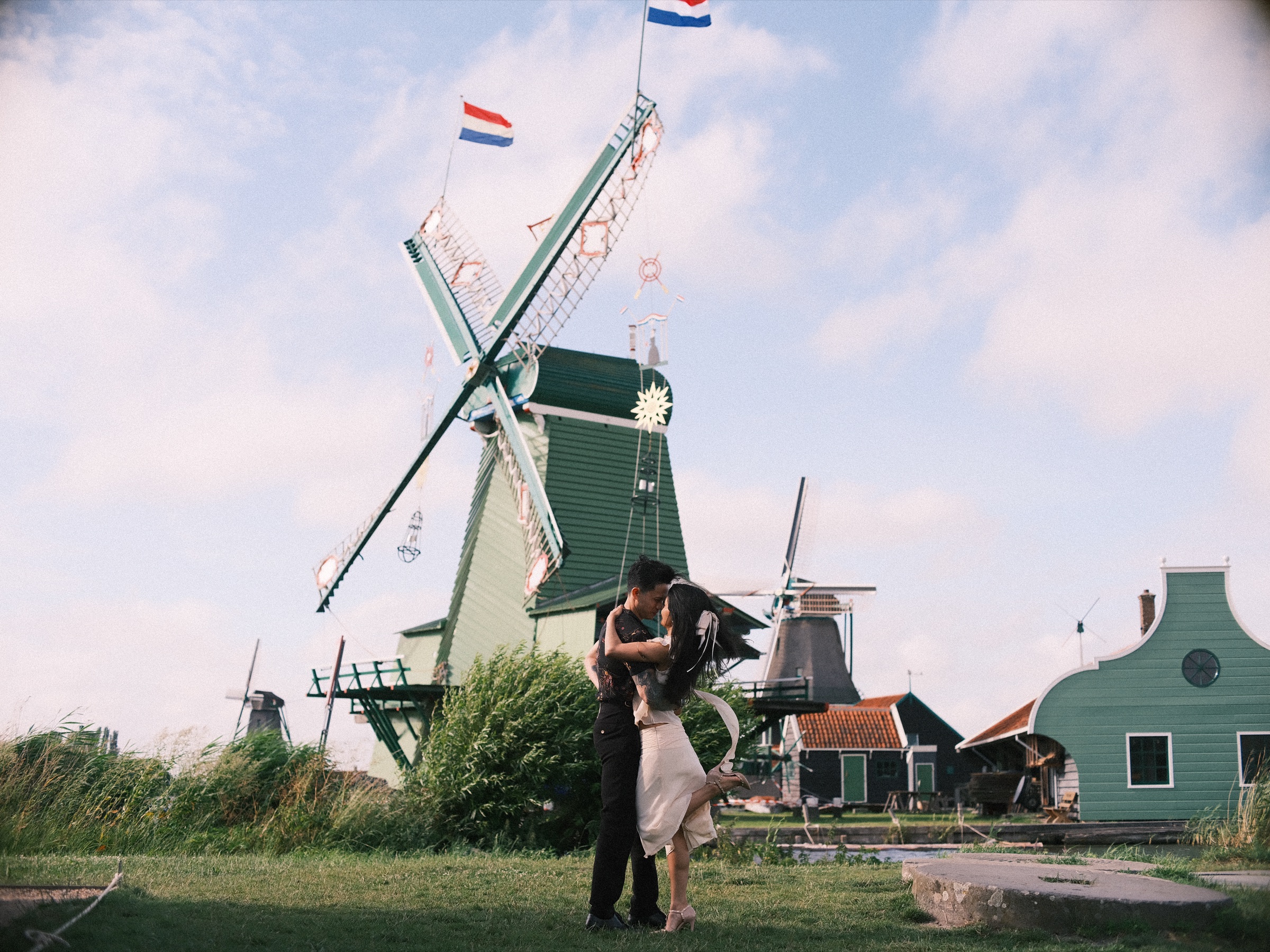 Man holding his partner close as she kicks one heel up, below a green mill flying Dutch flags with decorated sails, at the Zaanse Schans
