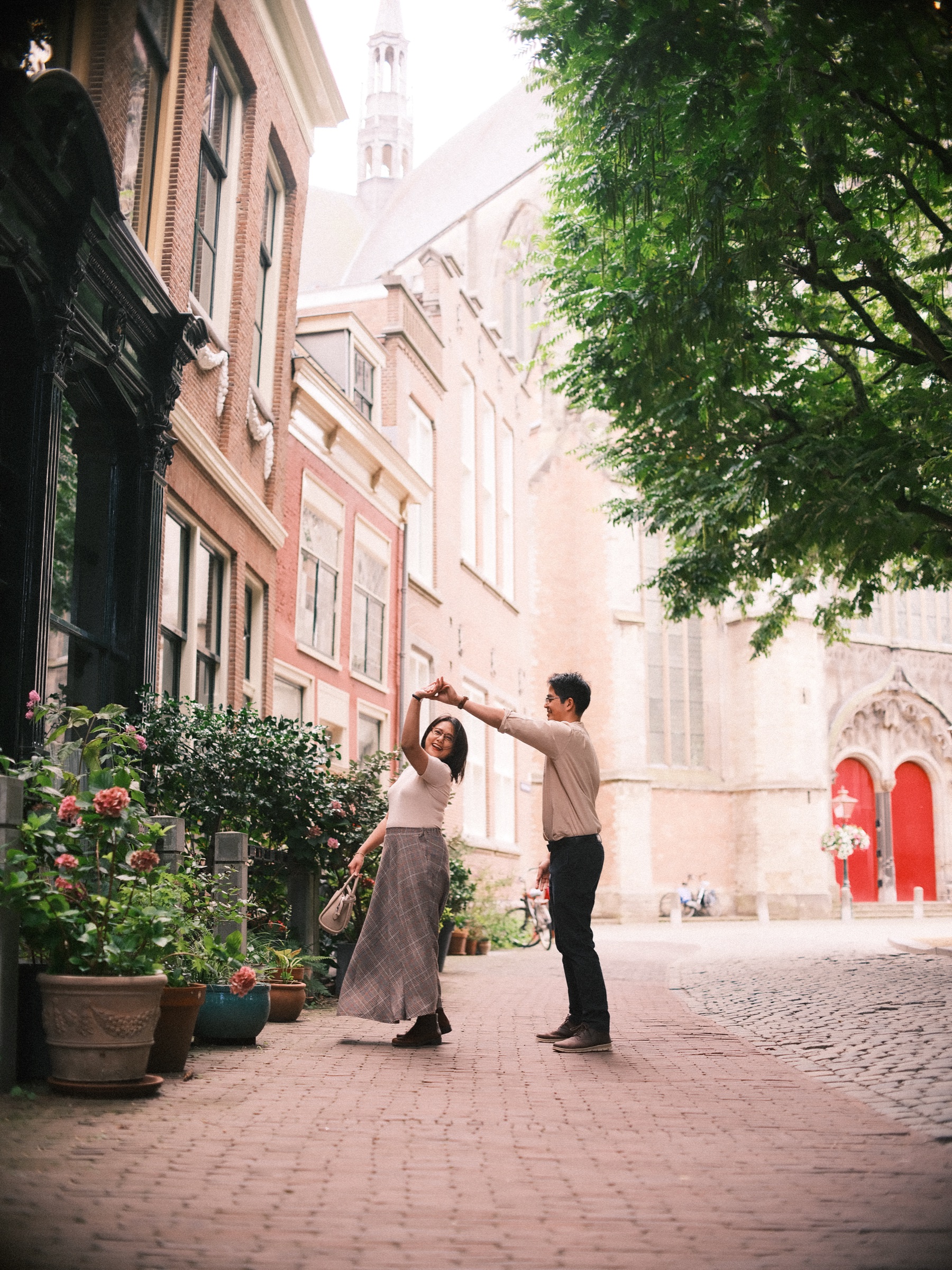 Couple dancing in a quiet street, his hand raised as she turns, potted roses beside them and the bright red doors of the Pieterskerk ahead