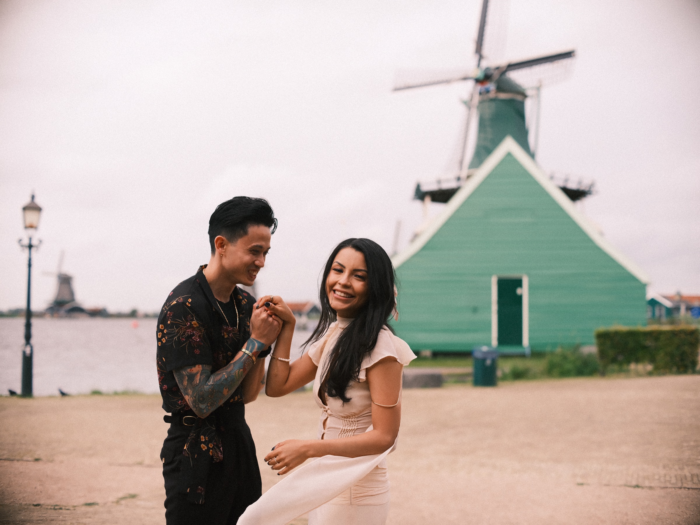 Couple dancing hand in hand on the quay at the Zaanse Schans, both laughing, a green wooden house and its windmill behind them