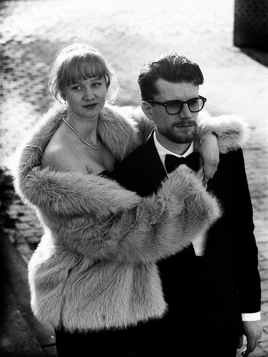 From above, a woman in a fur-collared coat embraces her partner in glasses and a bow tie in a close film portrait