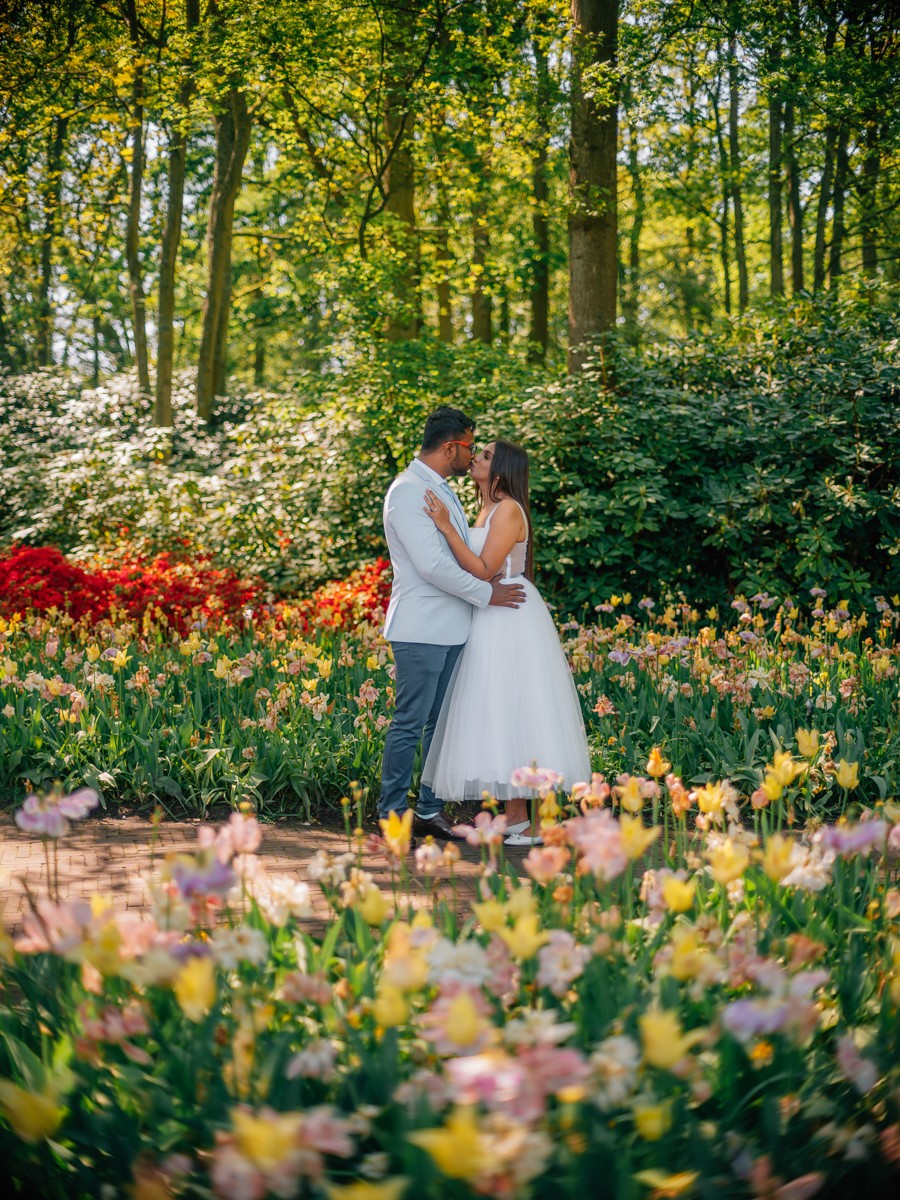 Bruidspaar omhelst elkaar onder hoge bomen, met perken vol gele, roze en rode tulpen in volle bloei om hen heen