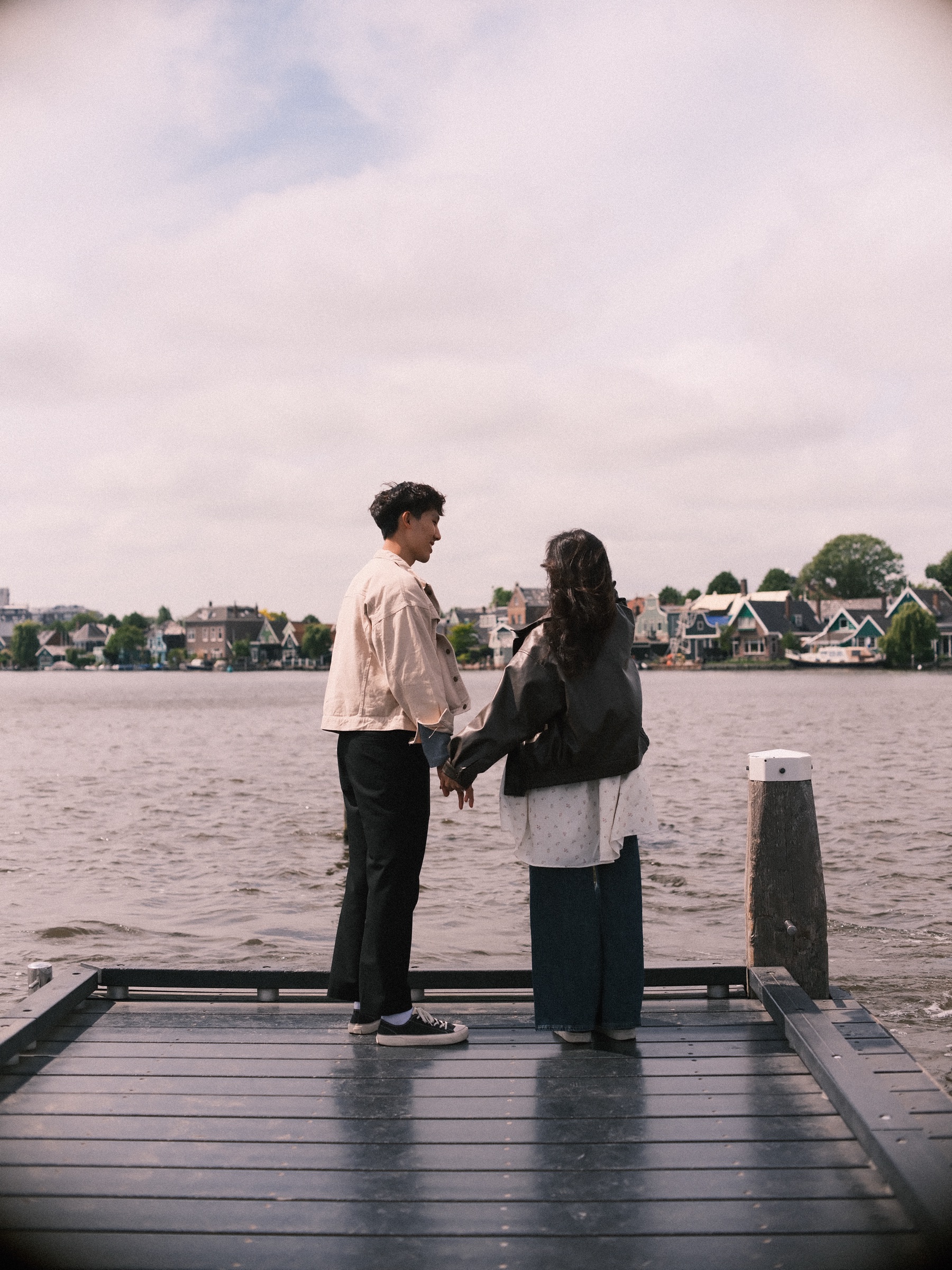 Back view of a couple holding hands at the end of a jetty, a wooden mooring post beside them and old houses on the far bank of the Zaan