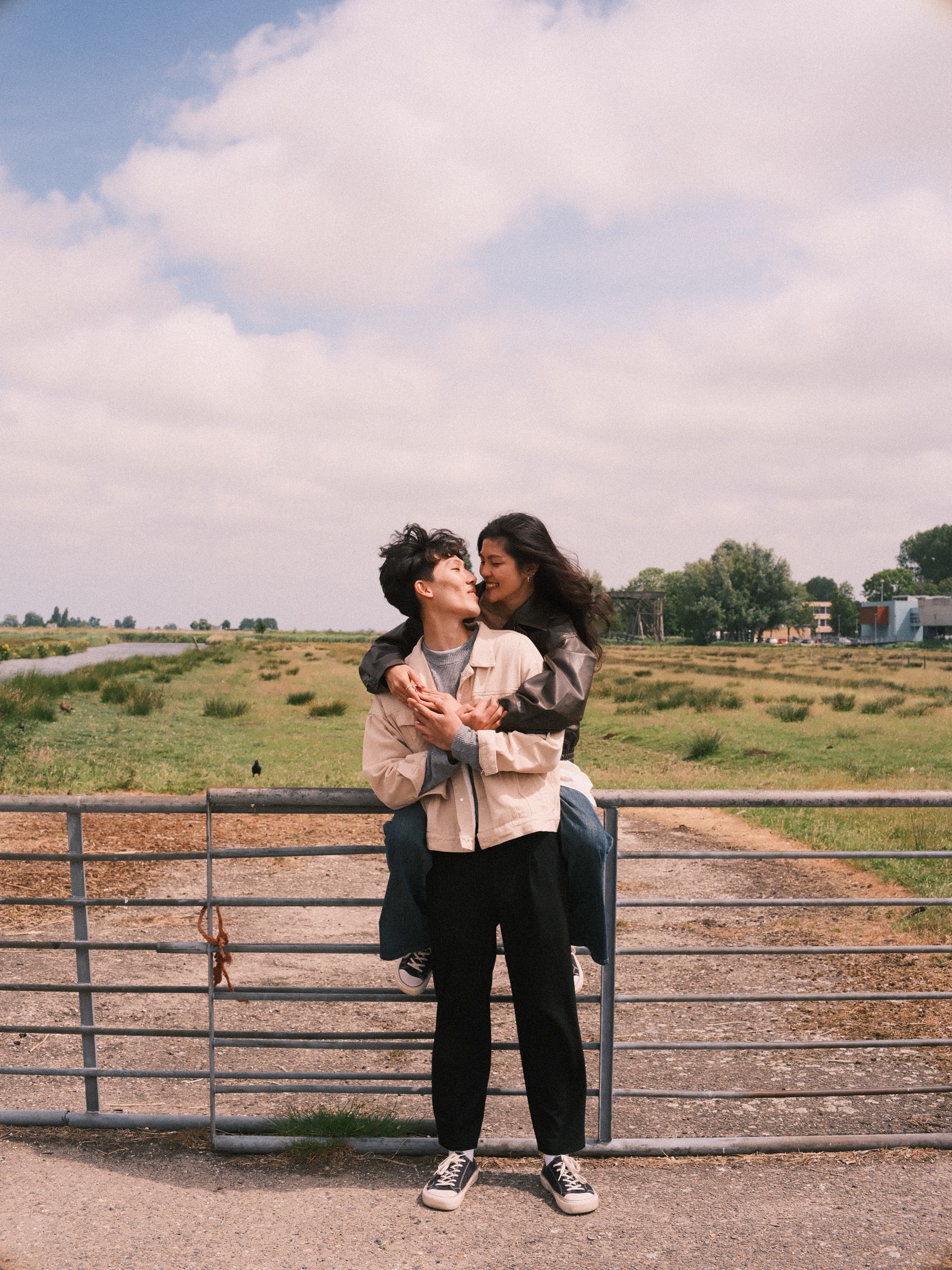 Woman hugging her partner from behind as she sits on a metal farm gate, flat green fields and a ditch stretching out under a cloudy sky
