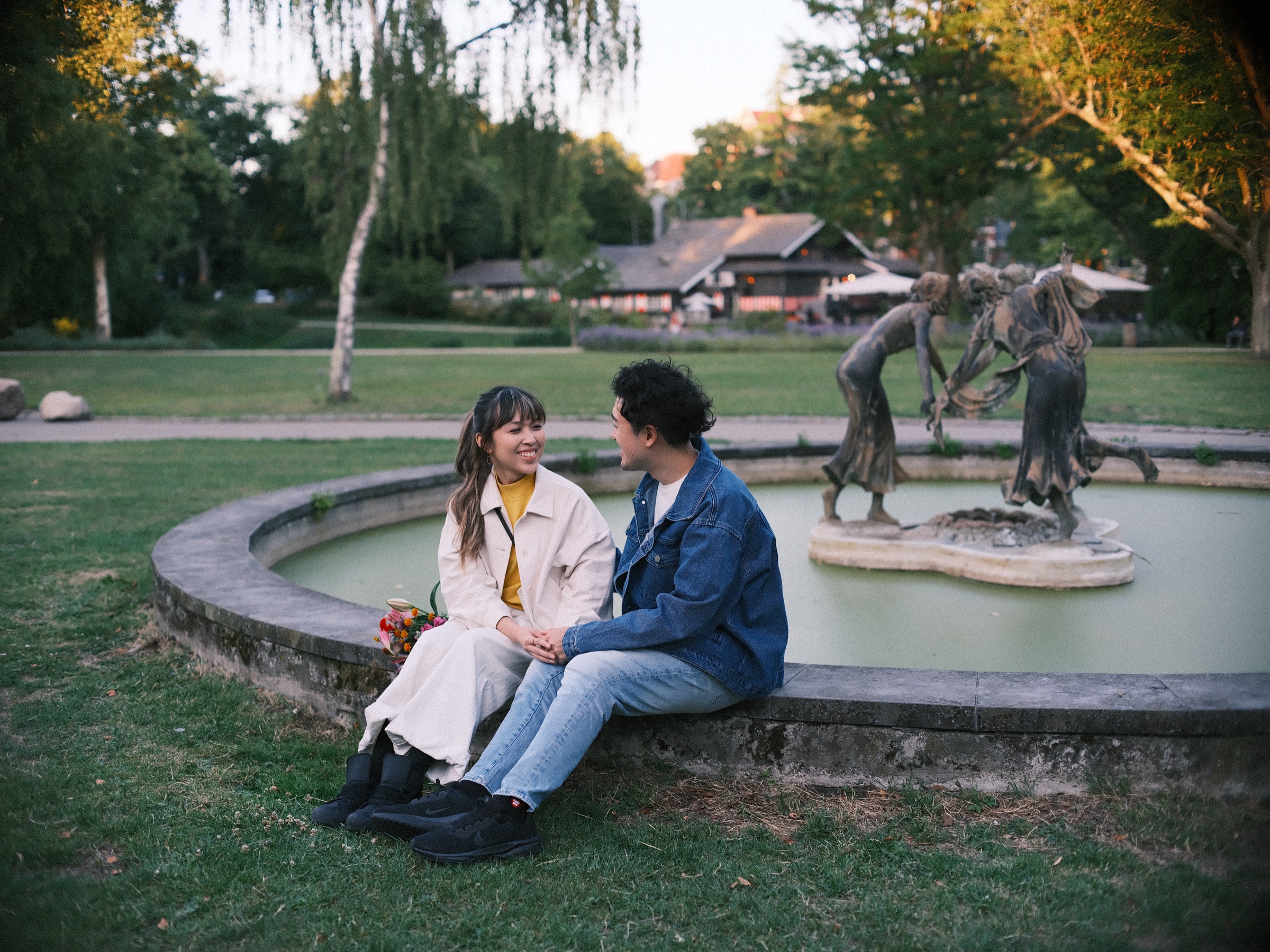 Stel op de rand van een ronde fontein met bronzen dansende figuren in Het Park, met een donker houten chalet en wilgen erachter