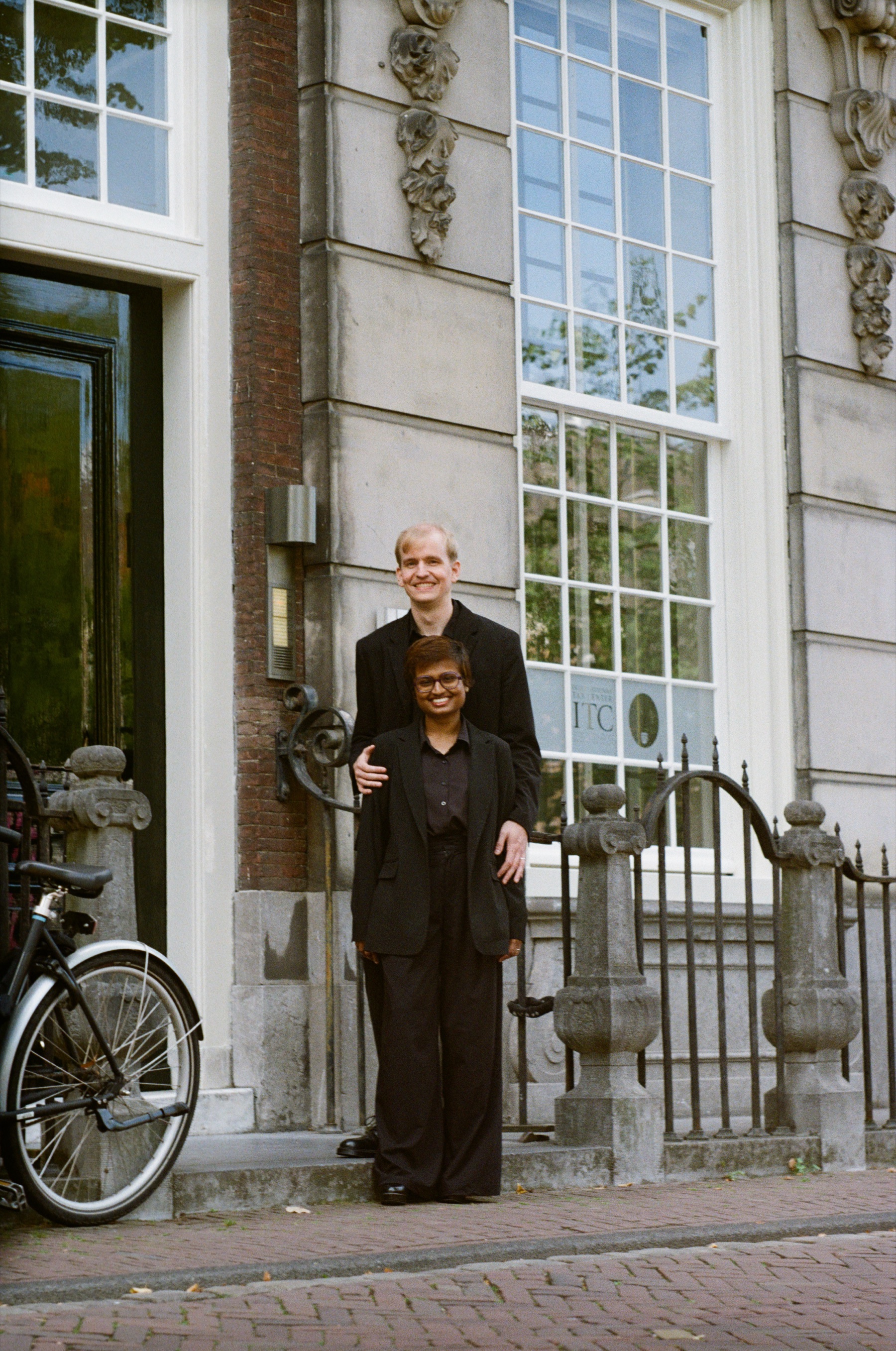 She stands in front with his hands on her shoulders, a grand stone facade with tall windows and carved ornaments behind them