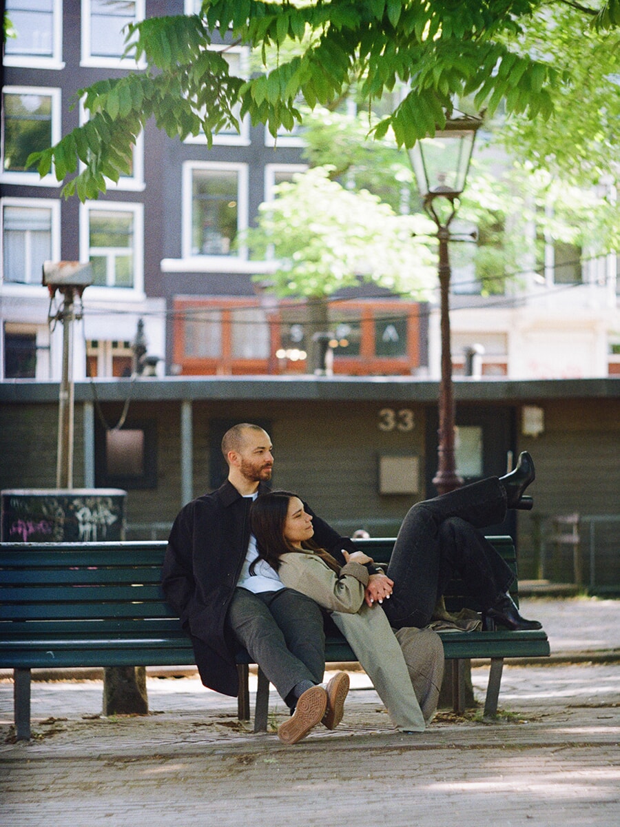 On a green canal-side bench in Amsterdam, a woman reclines against her partner with her boots raised in a colour film portrait