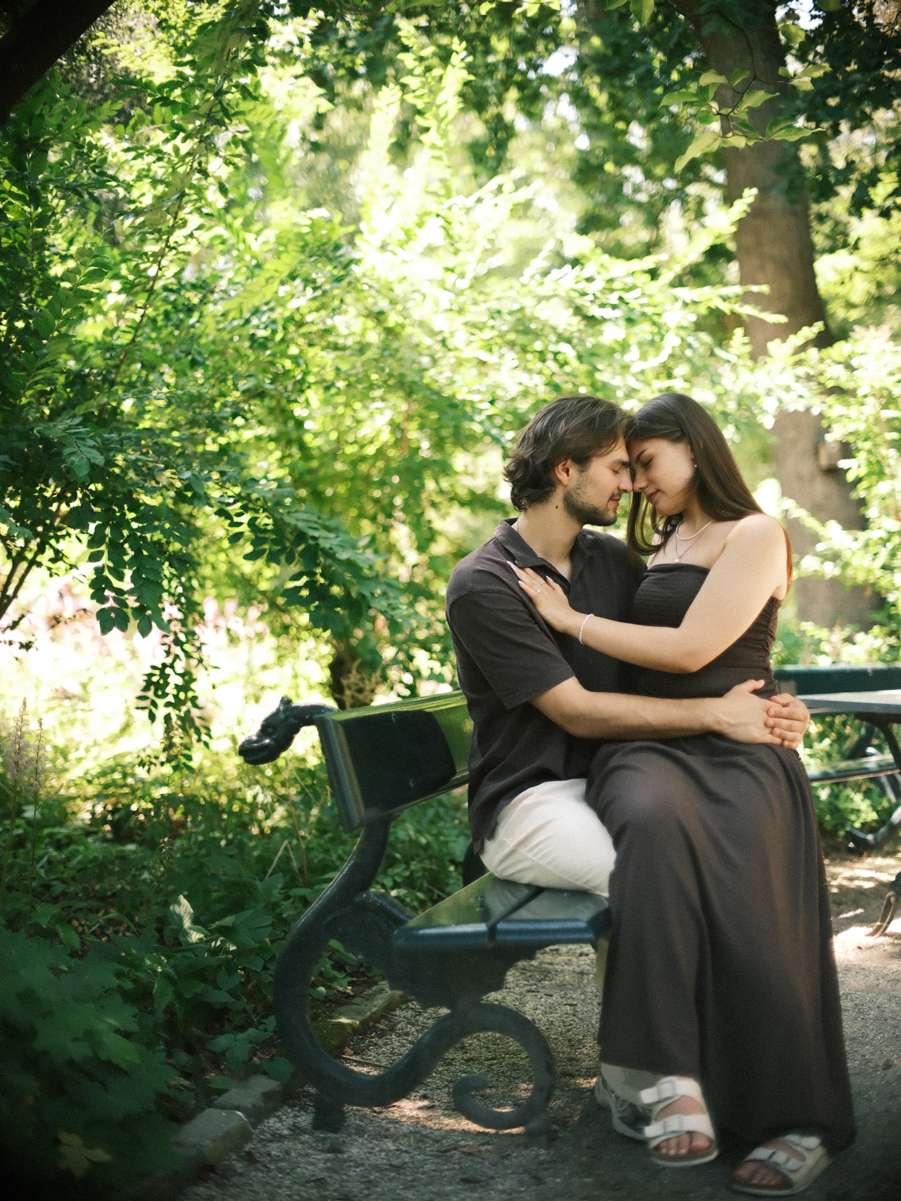 Couple on a green iron bench in leafy shade, she sits on his lap and they rest their foreheads together in a quiet corner of Trompenburg