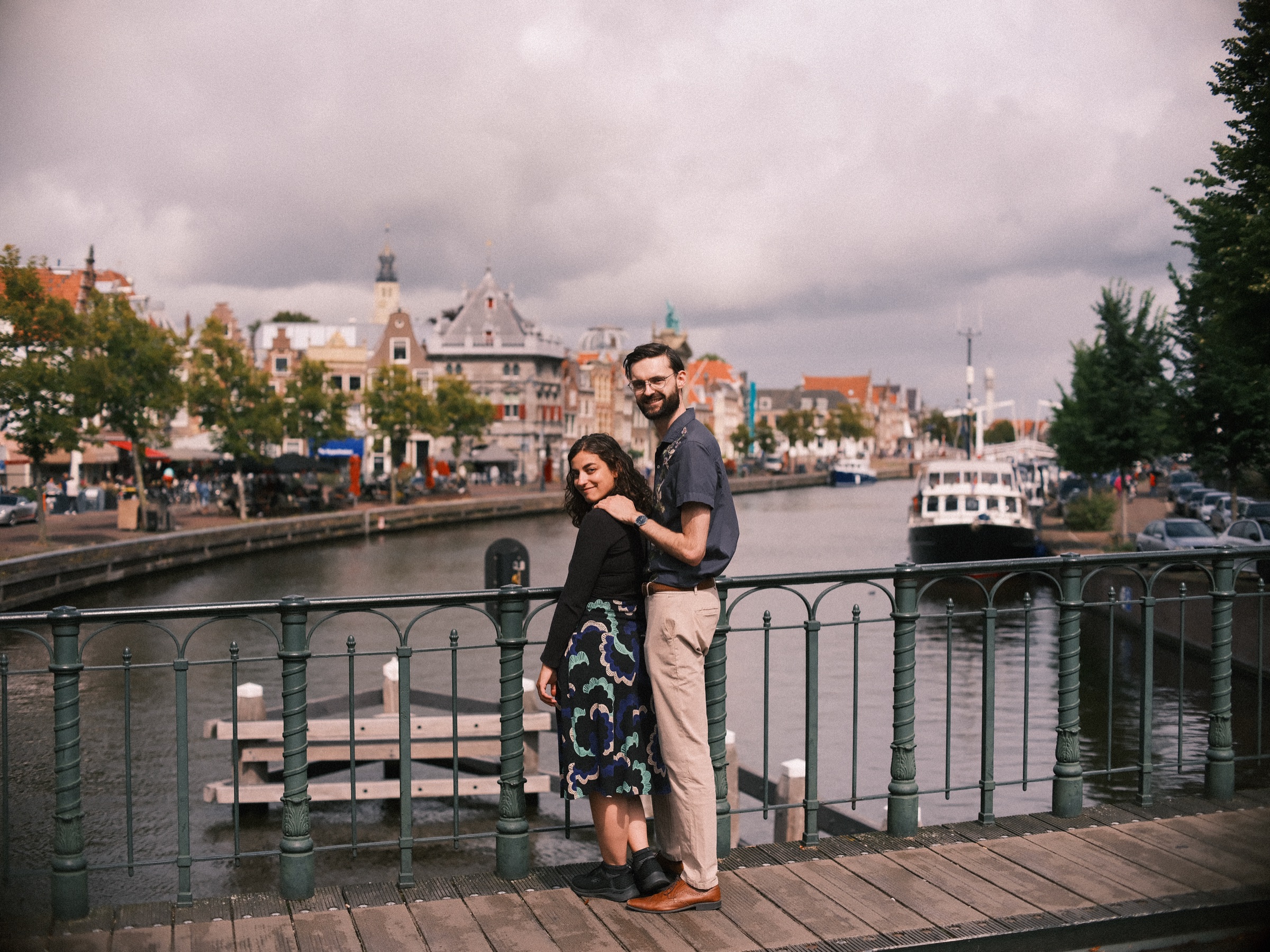 Stel bij de groene ijzeren leuning van een brug over het Spaarne in Haarlem, met sierlijke gevels en een witte boot onder een grijze lucht