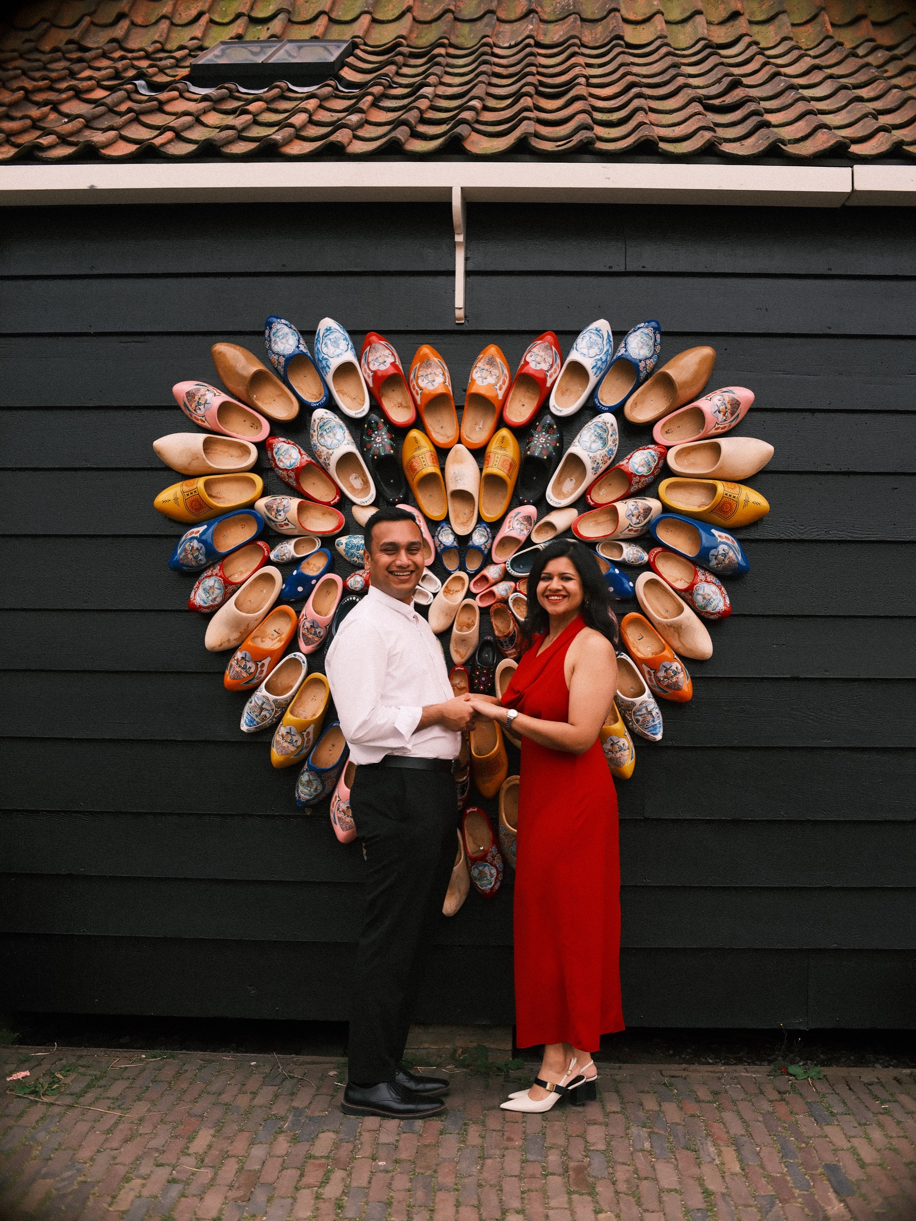 Couple holding hands in front of a big heart of painted wooden clogs on a dark wooden wall, she in a red dress and he in a white shirt