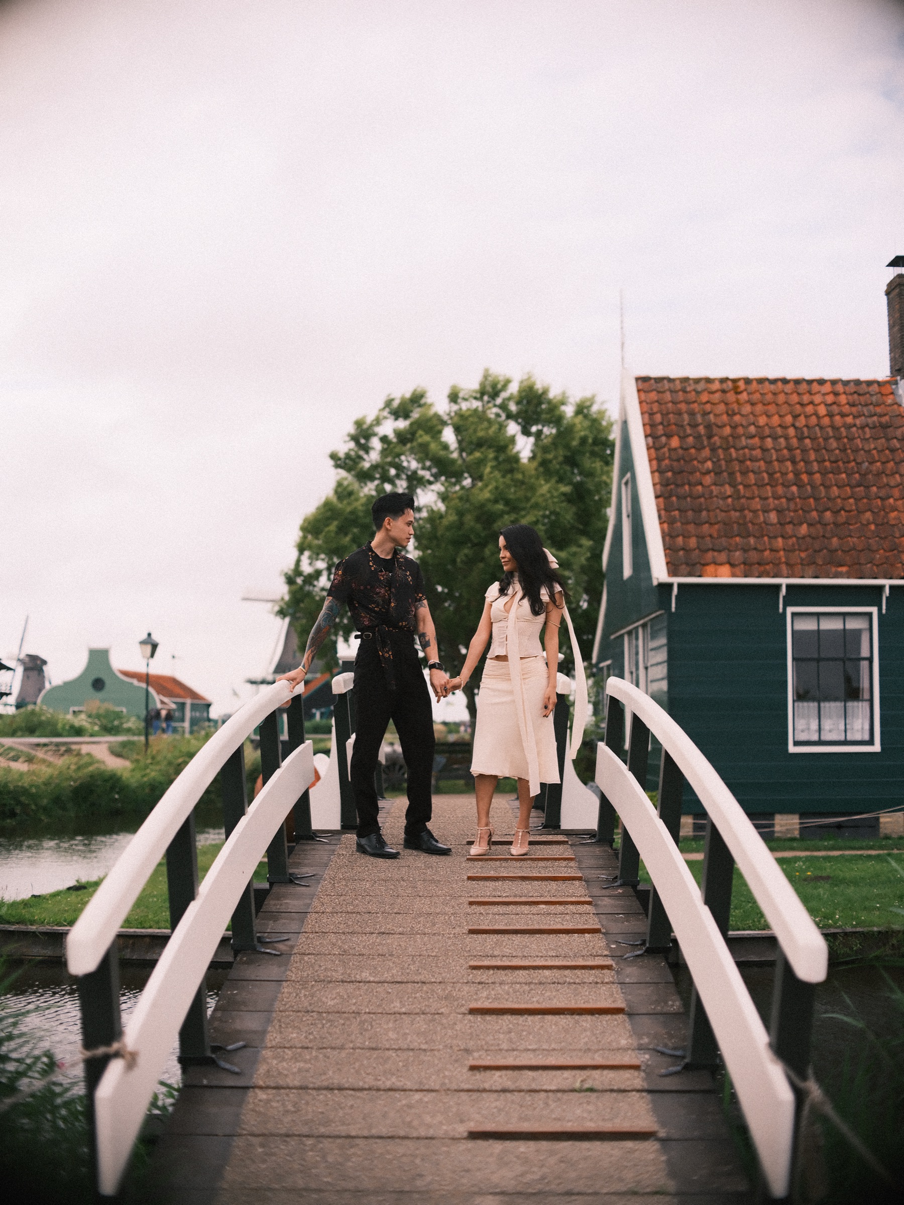 Couple holding hands on a white bridge in front of a green house with a red roof, the last frame of their Zaanse Schans session
