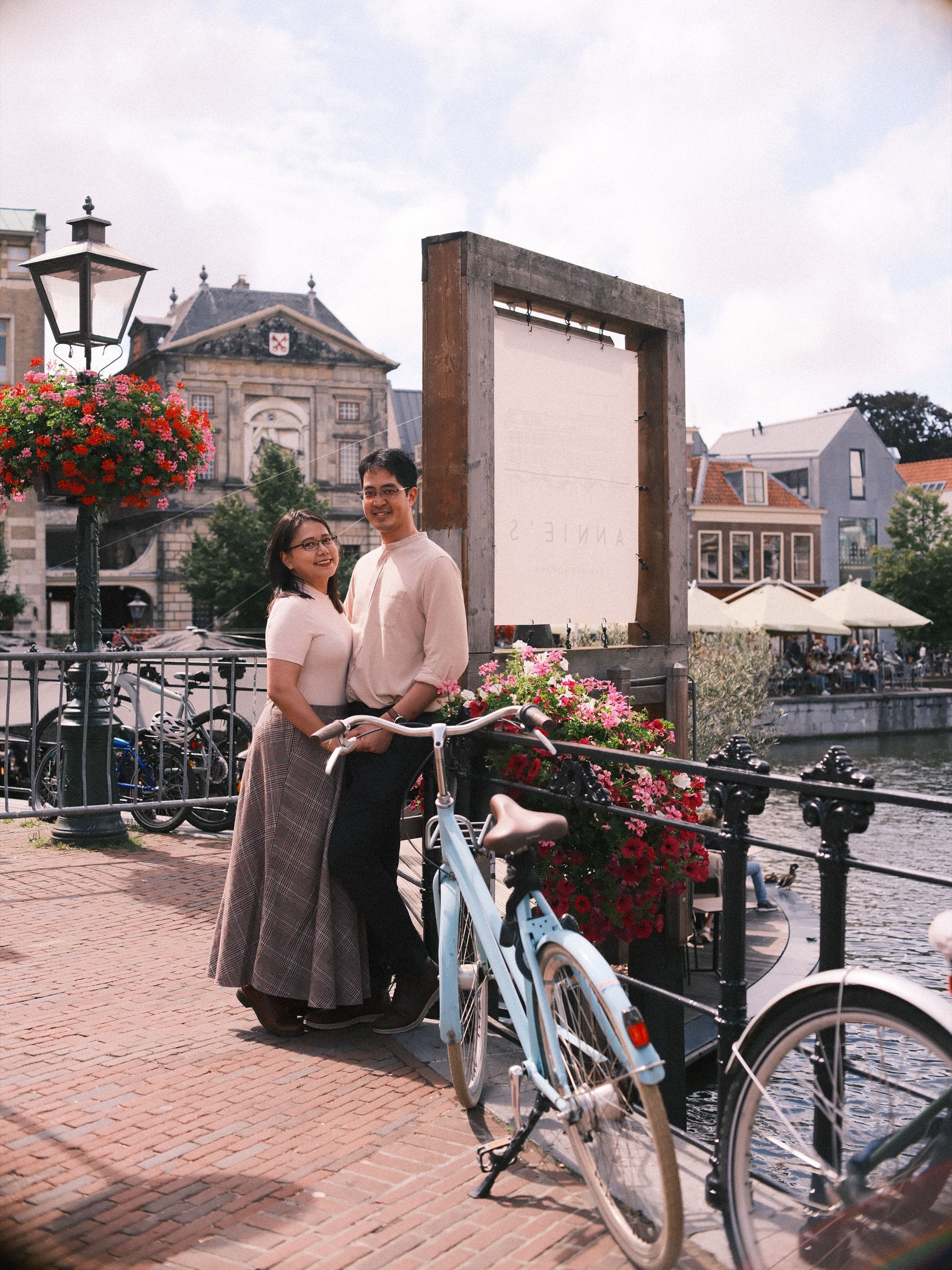 Couple by the railing of the Hoogstraat, a photo spot in Leiden with a lamppost full of red geraniums and the Waag across the water