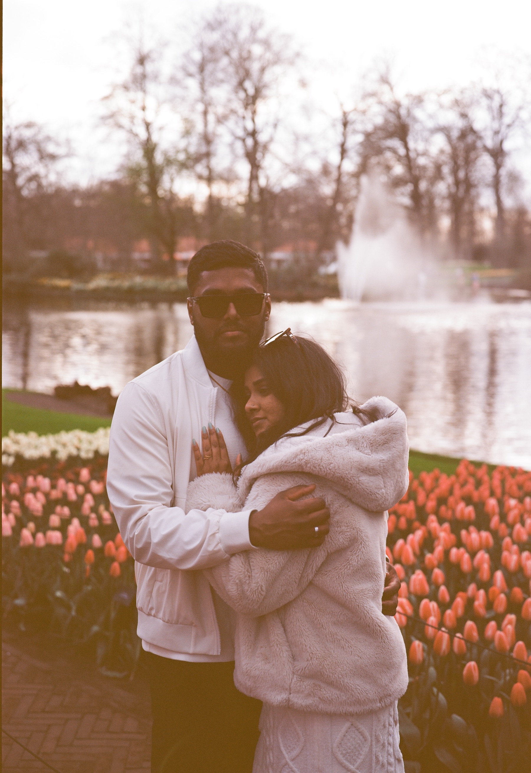 Woman resting against her fiancé’s chest beside a pond with a fountain, both dressed in white on a gray spring evening