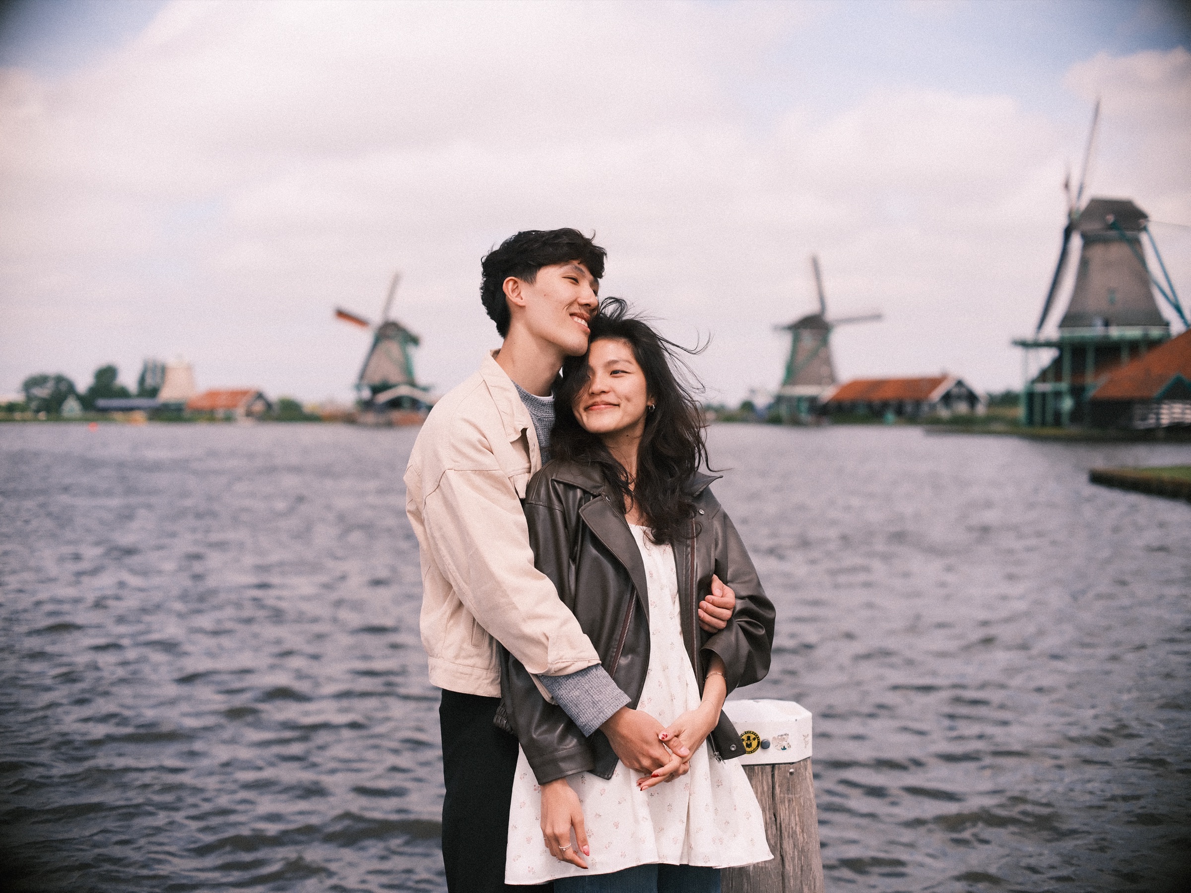 Man holding his fiancée from behind on a wooden jetty, the windmills of the Zaanse Schans lined up across the gray water of the Zaan